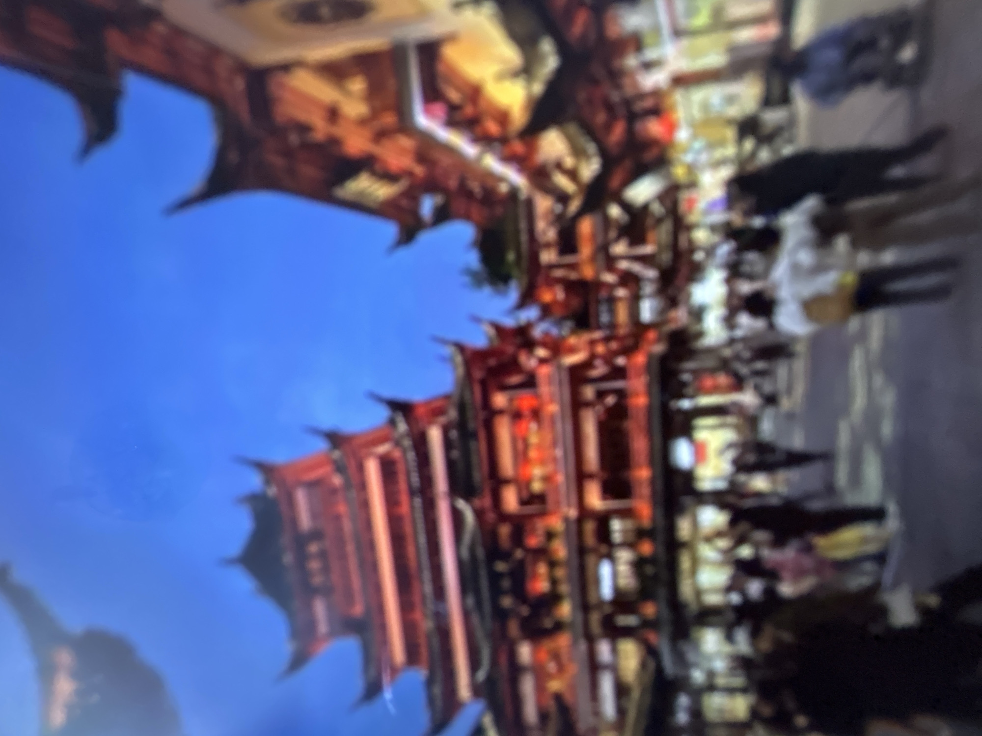 The image depicts a nighttime street scene in what appears to be a busy commercial area. The architecture suggests an East Asian influence, with traditional tiered roofs and ornate details on the buildings, illuminated by numerous red lanterns and lights. The blue sky suggests it is evening or night.
People are visible walking along the street, some singly and others in small groups. They are dressed in casual clothing. The overall atmosphere appears lively and bustling, consistent with a popular shopping or entertainment district at night. There is no discernible text in the image. The scene conveys a sense of vibrant nightlife and cultural ambiance.