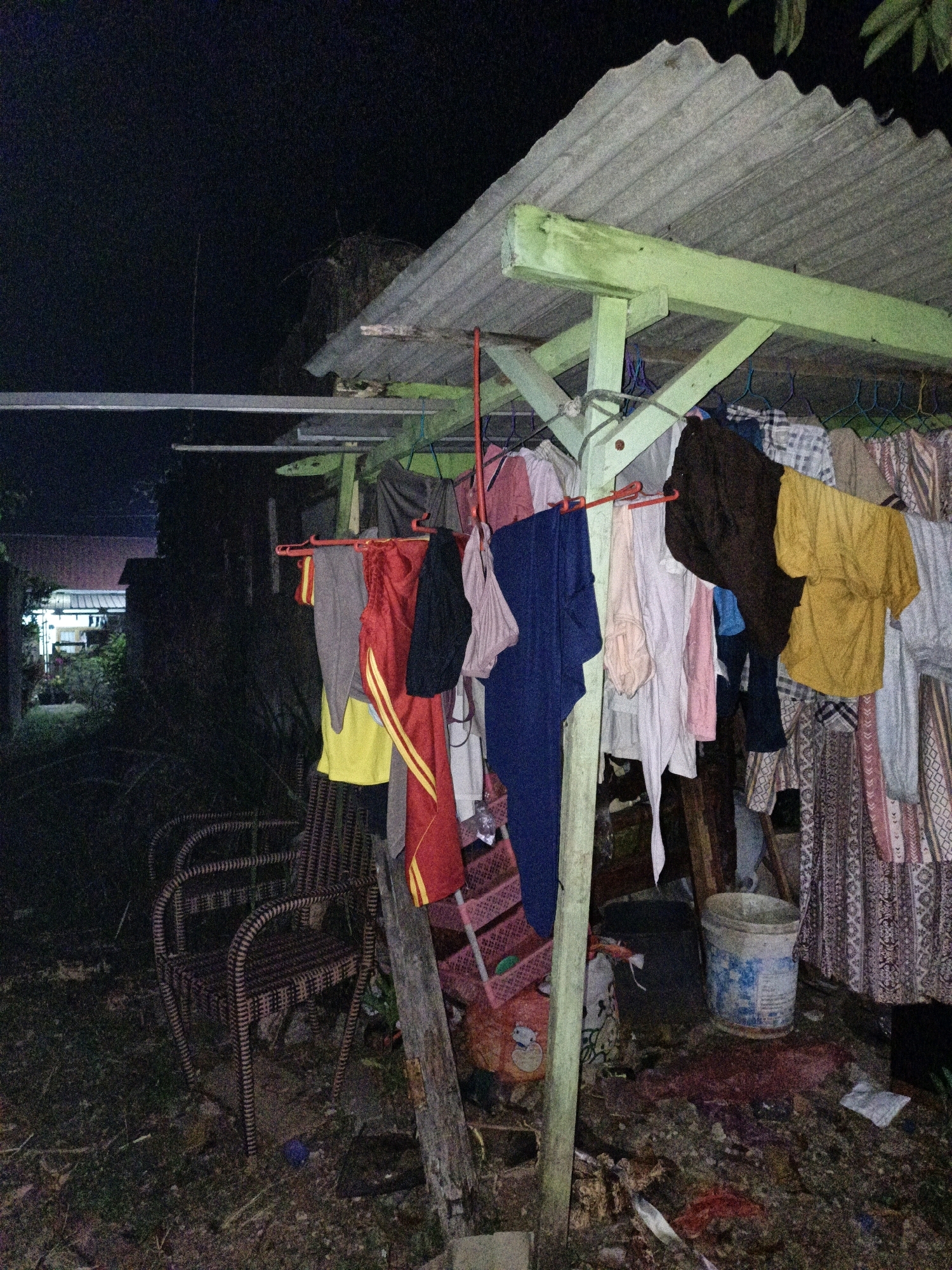 A low-angle, nighttime photograph depicts an outdoor laundry area adjacent to a modest dwelling in Cikal, Indonesia. The scene is cluttered with various items, including laundry hanging on a clothesline, a wicker chair, a bucket, and scattered debris on the ground. The corrugated metal roof of the laundry structure is supported by green wooden beams. The background reveals a dimly lit residential area with houses and vegetation. The overall impression is one of a rustic and somewhat unkempt environment.