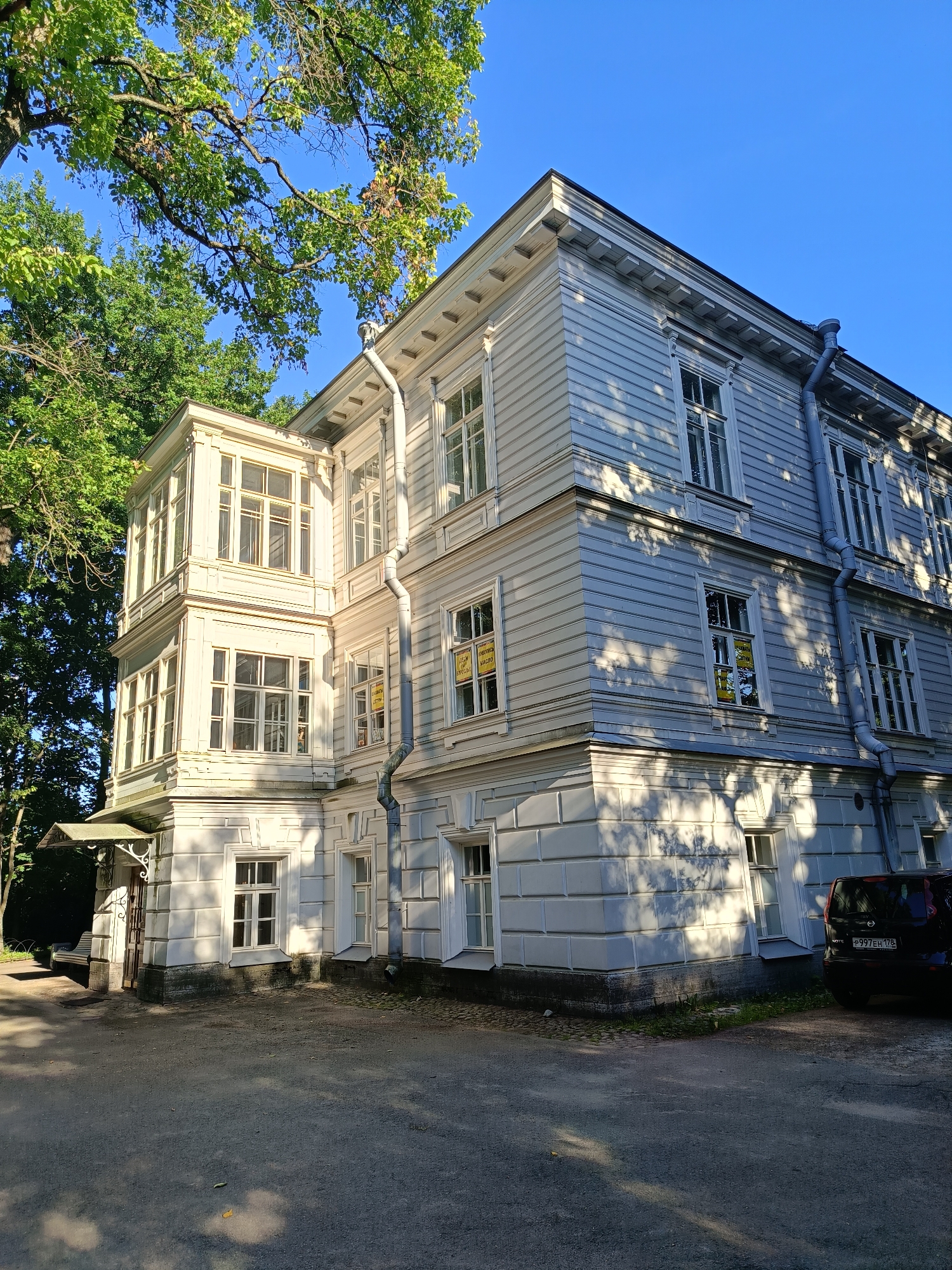 An exterior, eye-level, wide shot captures a white, two-story wooden building in a park-like setting. The building, featuring a bay window on the left and an attached garage or storage area on the right, is partially shaded by large trees. The building has a classic architectural style with horizontal siding, decorative trim, and multiple windows. Some of the windows have yellow signs posted in them. The ground in front of the building is a paved driveway or walkway. A dark-colored car is parked on the right side of the image. The sky is clear and blue.
