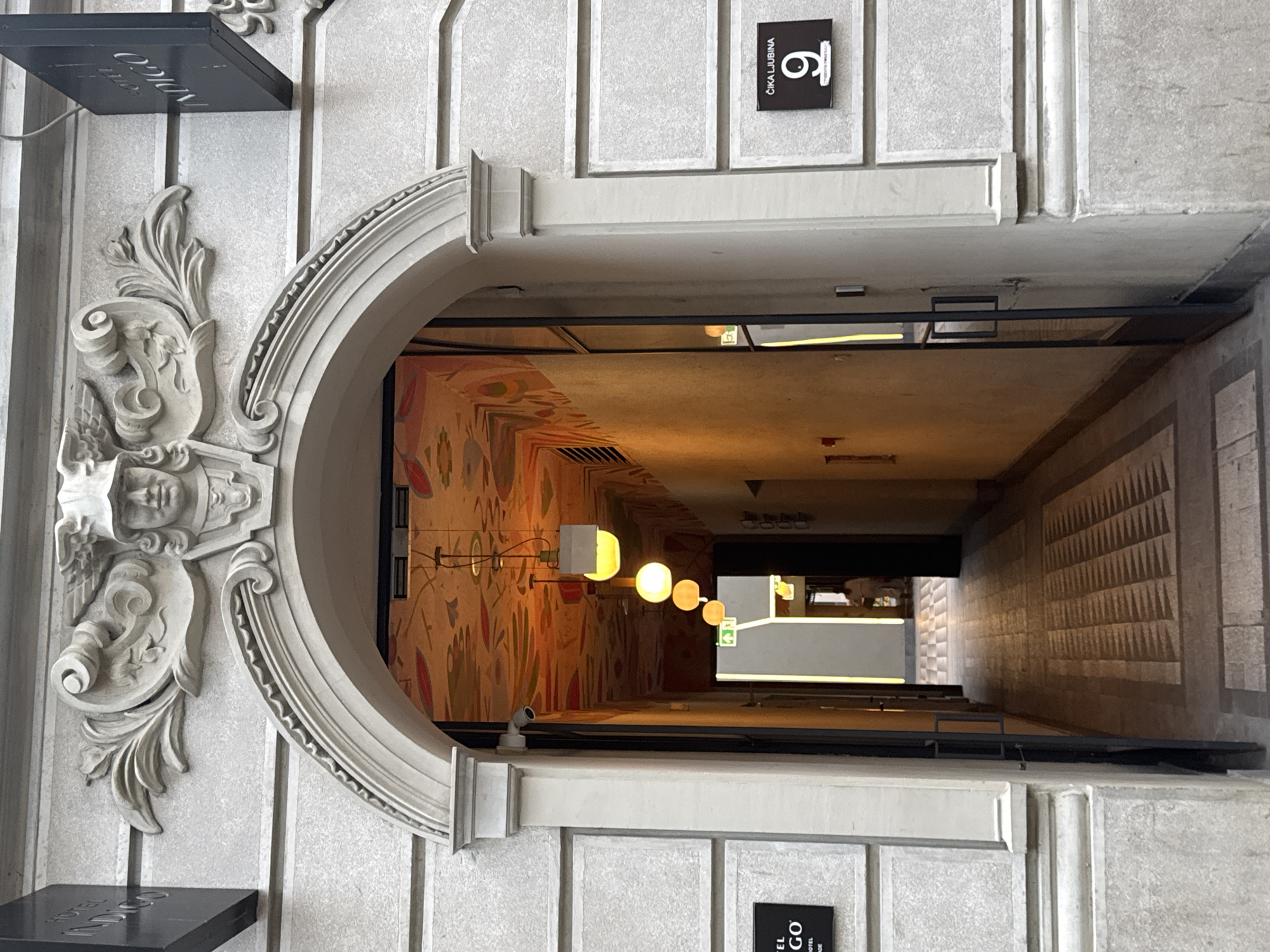 The image shows the entrance to a building in Belgrade, Serbia, identified by the street sign "Čika Ljubina 9" which is located on the right side of the building. The building's facade is made of light-colored stone with decorative architectural elements above the arched entryway. Above the arch, a relief sculpture depicts a face wearing a helmet and flanking foliage.
A black sign reading "HOTEL INDIGO" is mounted to the left of the entrance, and another identical sign is visible to the right. Through the open archway, a dimly lit corridor is visible, leading into the building. The corridor's ceiling is decorated with a vibrant, colorful mural. Several pendant lights, emitting a warm yellow glow, are suspended from the ceiling in the corridor. The floor of the corridor is paved with patterned tiles. A security camera is mounted on the wall to the left of the entrance. Further down the corridor, a person can be seen in the distance, standing near an exit or another doorway. The overall impression is of a historic building with modern interior design elements.
