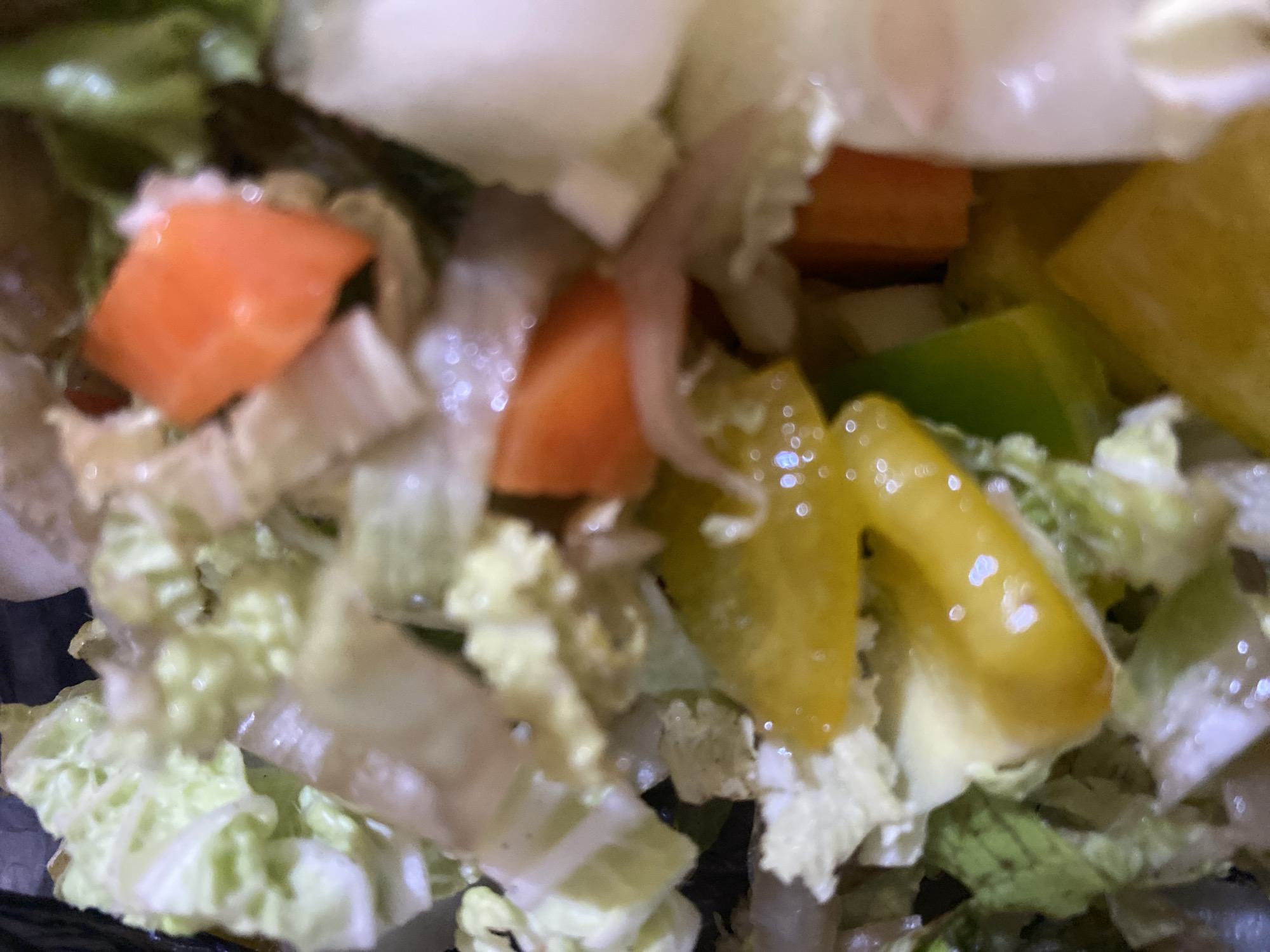The image shows a close-up of a chopped salad. The salad contains leafy greens, diced carrots, and yellow and green bell peppers. The ingredients are mixed together in a bowl, which is dark in color and not fully visible. The lighting creates some glare and highlights on the vegetables. There is no indication of the city of Bar Jirgi Summa, Nigeria, in the image.