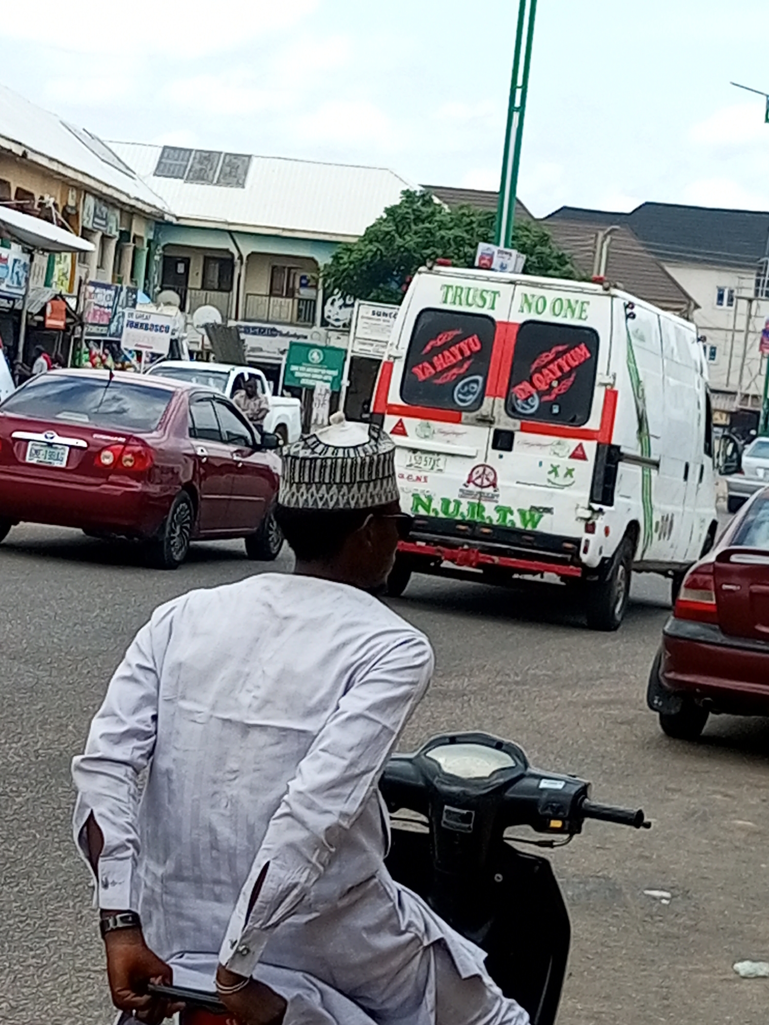 The image depicts a street scene in Arawa, Nigeria, likely on a cloudy day. In the foreground, a man wearing a light-colored, long-sleeved traditional outfit and a patterned cap is seated on a dark-colored scooter, facing away from the camera. His hands are on the scooter's handlebars. Behind him, a white van with red accents is visible. The rear of the van has the words "TRUST NO ONE" emblazoned across the top. Below this, two red-bordered windows contain text and graphics, with "YA HAYYU" on the left and "QAYYUM" on the right, and what appear to be smiley face emojis. The van also displays "N.U.R.T.W." in green letters on its lower rear. To the left of the van, a red sedan is parked, facing the same direction as the van, with its license plate visible. Further back, other vehicles and buildings line the street, suggesting a commercial or residential area. Signs are visible on the buildings, some with writing that is difficult to decipher due to image resolution. The road surface appears to be asphalt.