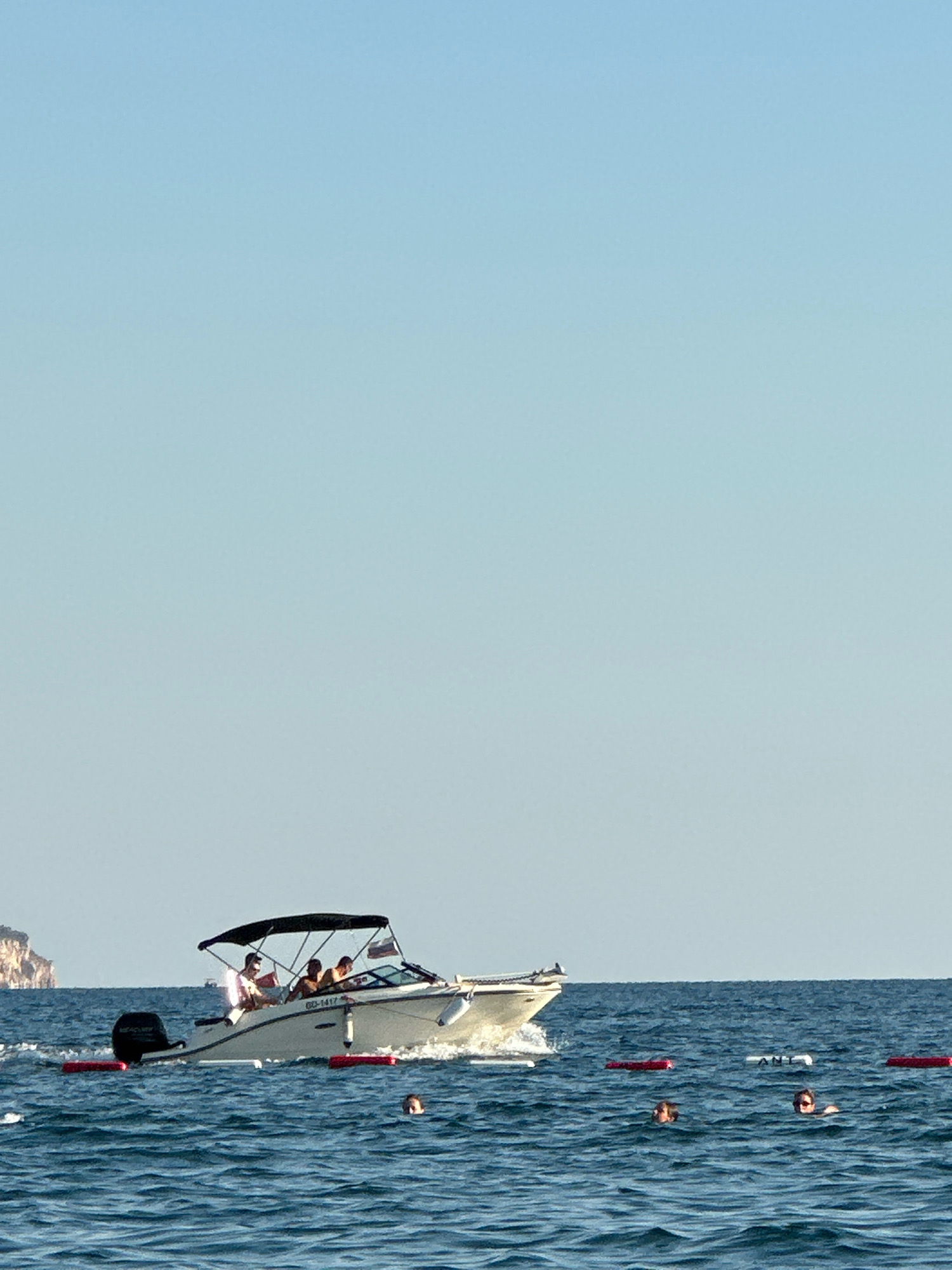 A white motorboat with a black canopy is moving through blue ocean water under a clear sky. The boat has at least three people visible onboard. In the foreground, several people are swimming in the water. Red buoys are floating in the water, indicating a swimming area or hazard. In the background, a rocky, green-covered cliff is visible. The overall scene suggests a sunny day at a coastal location, possibly the coast of Boreti, Montenegro, given the distinctive rocky cliff formation which is characteristic of the region.