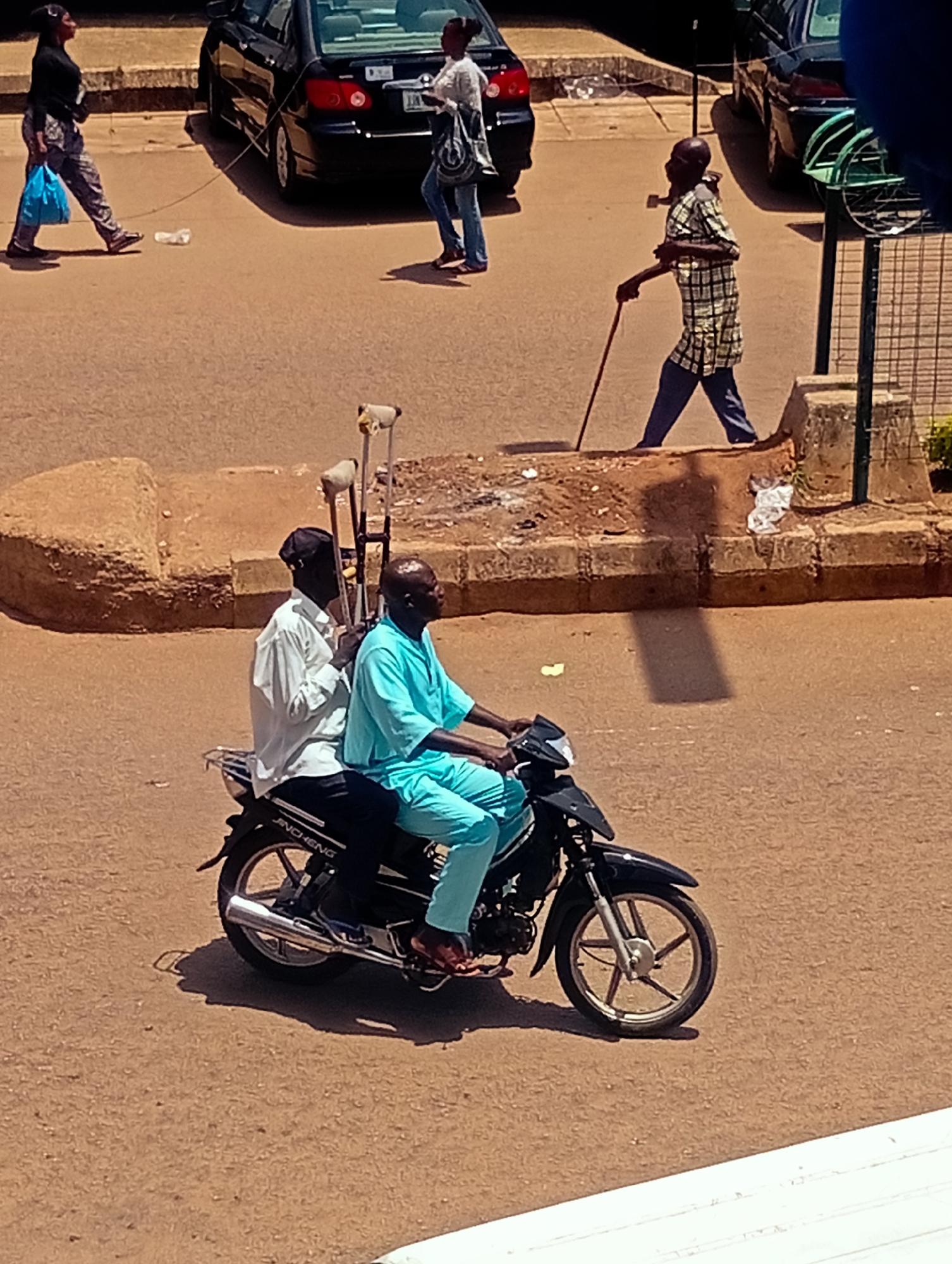In Bukuru, Nigeria, two individuals are riding a black motorcycle. The passenger in the back is wearing a white long-sleeved shirt and a black cap. The driver in the front is wearing a light blue tunic. Two crutches are attached to the back of the motorcycle. In the background, a man in a checkered shirt and blue pants walks with a cane. Further in the distance, two women are walking on the sidewalk. One woman, wearing a black shirt and patterned pants, carries a blue bag and a plastic bottle on the ground is near her. The other woman, in a white shirt and blue jeans, carries a shoulder bag. Several cars are parked on the street, including a black sedan. The ground is a dirt or gravel road, and there is a raised concrete curb. The image is taken from a high vantage point.