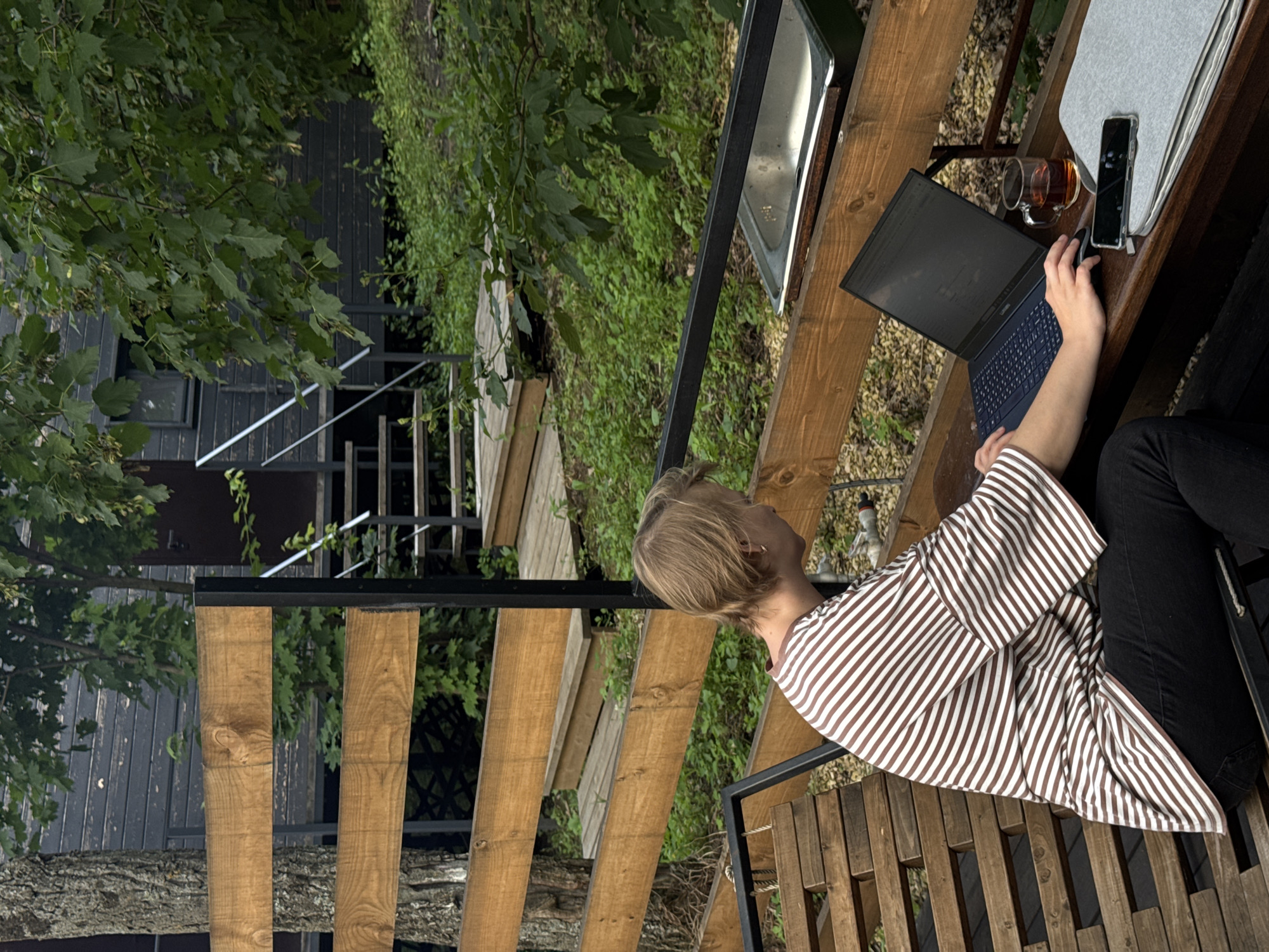 A person with short blonde hair, wearing a brown and white striped shirt, sits at a wooden table outdoors, working on a laptop. A glass of amber liquid, likely tea, sits next to the laptop, along with a smartphone and a light gray notebook. The scene appears to be on a wooden deck or balcony, with a wooden railing and steps visible. In the background, lush green trees and foliage suggest a natural or wooded setting. A dark-colored building with a window is partially visible through the trees. The overall lighting suggests it is daytime. The person's posture and focus on the laptop indicate they are engaged in work or study. The location is identified as Ust-Kurdyum, Russia.