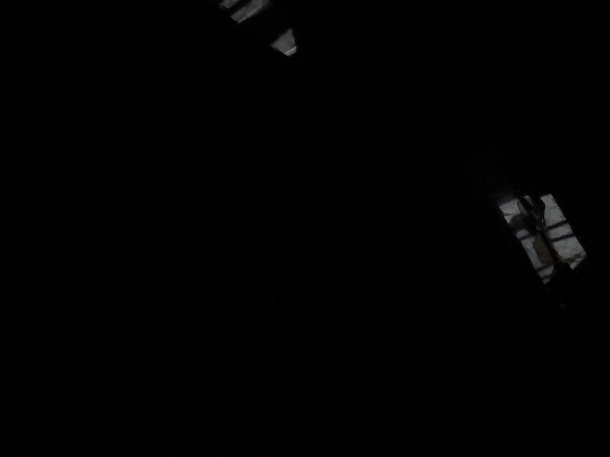 The image is a low-light, black and white photograph with the majority of the frame dominated by darkness. In the upper right quadrant, a sliver of what appears to be a light-colored structure or object is visible, possibly a roof or a piece of debris. In the lower portion of the image, more detailed shapes emerge from the darkness, showing what seem to be architectural elements. These include what could be window frames, beams, or corrugated material. The overall impression is of an interior or exterior space that is poorly illuminated, possibly abandoned or under construction. No people or discernible actions are present. The scene is set in an indeterminate location within a city, with no specific landmarks or identifying features visible.