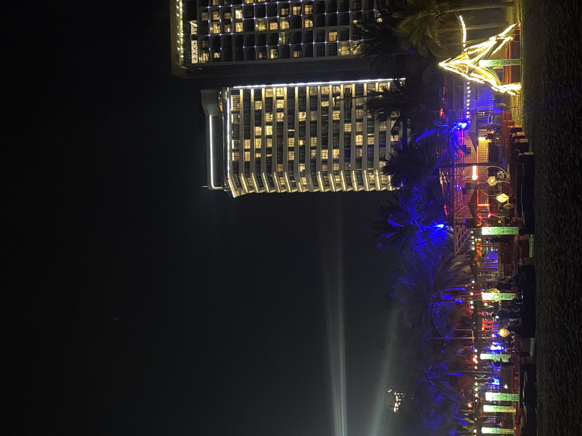 The image depicts a nighttime scene on a beach, likely in Đà Nẵng, Vietnam, given the architectural style of the buildings and the presence of palm trees. In the background, two modern high-rise buildings are illuminated, with many windows lit, suggesting they are hotels or residential complexes. The building on the left is a multi-story structure with a distinctive curved facade and white accent lighting along its edges. The building on the right is taller and appears more rectangular, with a grid-like pattern of lit windows.
In the foreground, a beach area is set up with various lights and seating arrangements, indicating an outdoor entertainment venue or event. Several tall, green cylindrical lights stand vertically on the sand. Scattered seating, possibly lounge chairs or cushions, are arranged in front of these lights and other structures. There are also decorative elements, including a triangular tent-like structure illuminated with golden fabric and lights, and circular light fixtures hanging in the distance. Palm trees are visible throughout the scene, some illuminated with blue and red lights, adding to the festive atmosphere. The sky is dark, suggesting it is late evening or night, with some faint white light beams visible in the upper left portion of the image, possibly from a projector or searchlight. A few silhouetted figures can be seen in the illuminated area, engaged in social activities. The sand of the beach is visible in the immediate foreground.