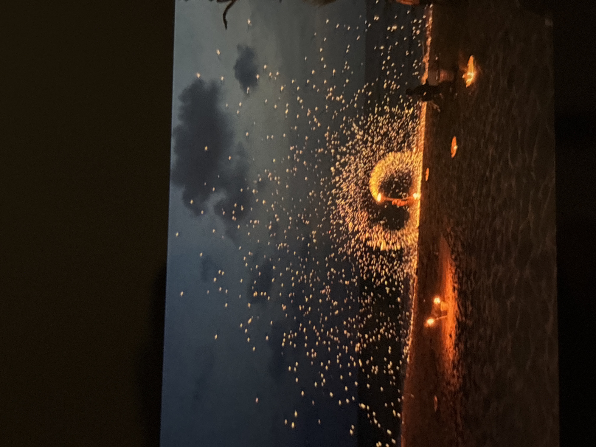 The image depicts a fire performance on a beach at dusk, likely in Chalong, Thailand. A performer is seen twirling a flaming object, creating a shower of sparks that arch across the scene. The dark sky is dotted with these glowing embers, some of which appear to be falling like raindrops. Several small, lit objects, possibly floating lanterns or candles, are scattered on the sand in the foreground, adding to the ambiance. The beach sand is visible, with the dark sea in the background. Silhouettes of a few people are discernible, observing the performance. The overall scene is illuminated by the fire and the glowing embers, creating a dramatic and ethereal effect against the darkening sky.