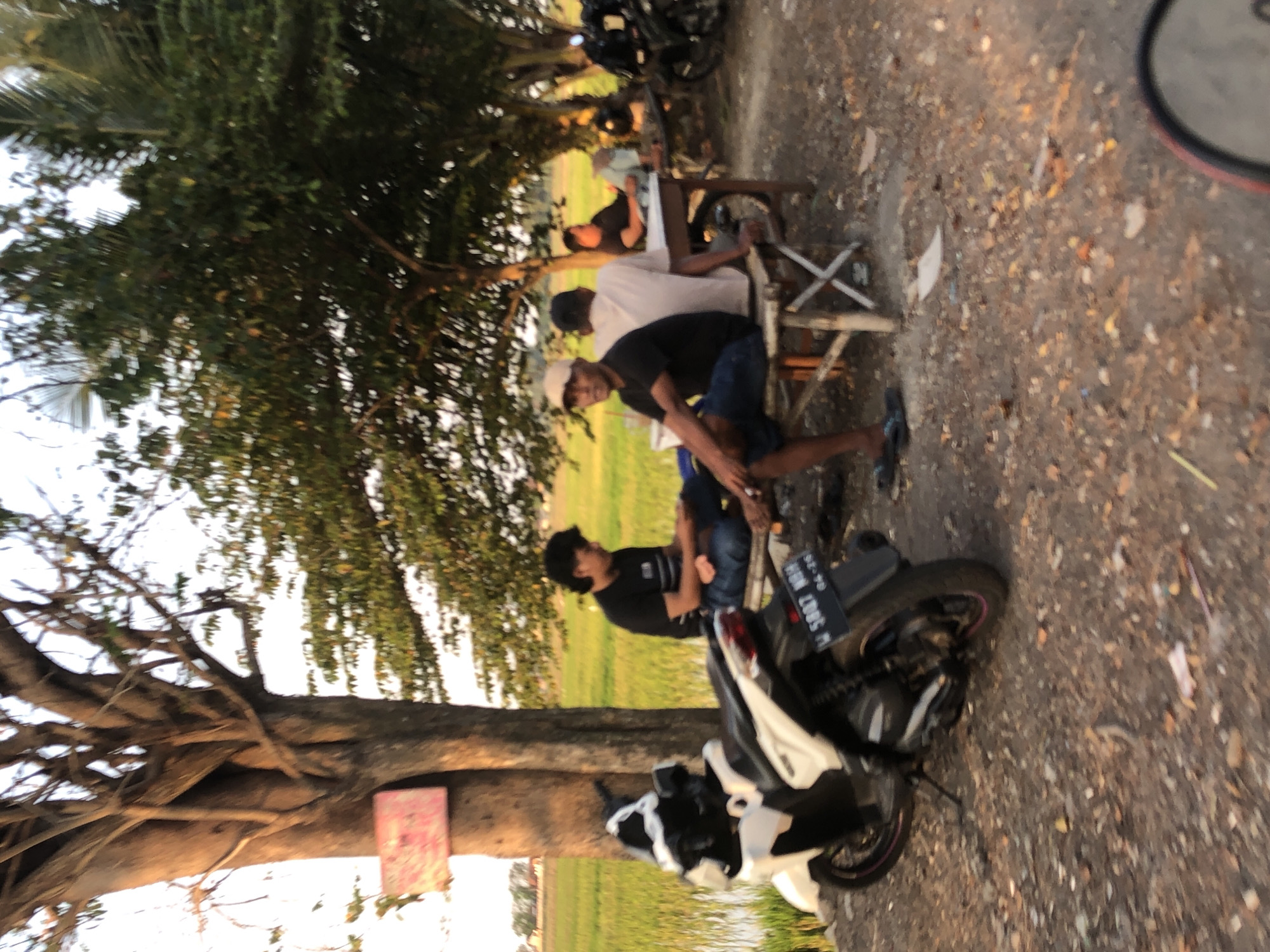 In the city of Keboguyang, Indonesia, on a dirt road bordering a rice paddy, several men are gathered under a large tree. A white scooter is parked on the left, its license plate reading "W 5887 NOH". A group of men are seated on a low bamboo table and benches. One man, wearing a black t-shirt and blue jeans, is facing the camera. Another man in a white t-shirt and blue jeans is seated with his back to the camera, while a third man, in a black t-shirt, sits further away. In the background, a rice field stretches out, and other trees dot the landscape. The sun is low in the sky, casting long shadows and creating a warm, golden light.