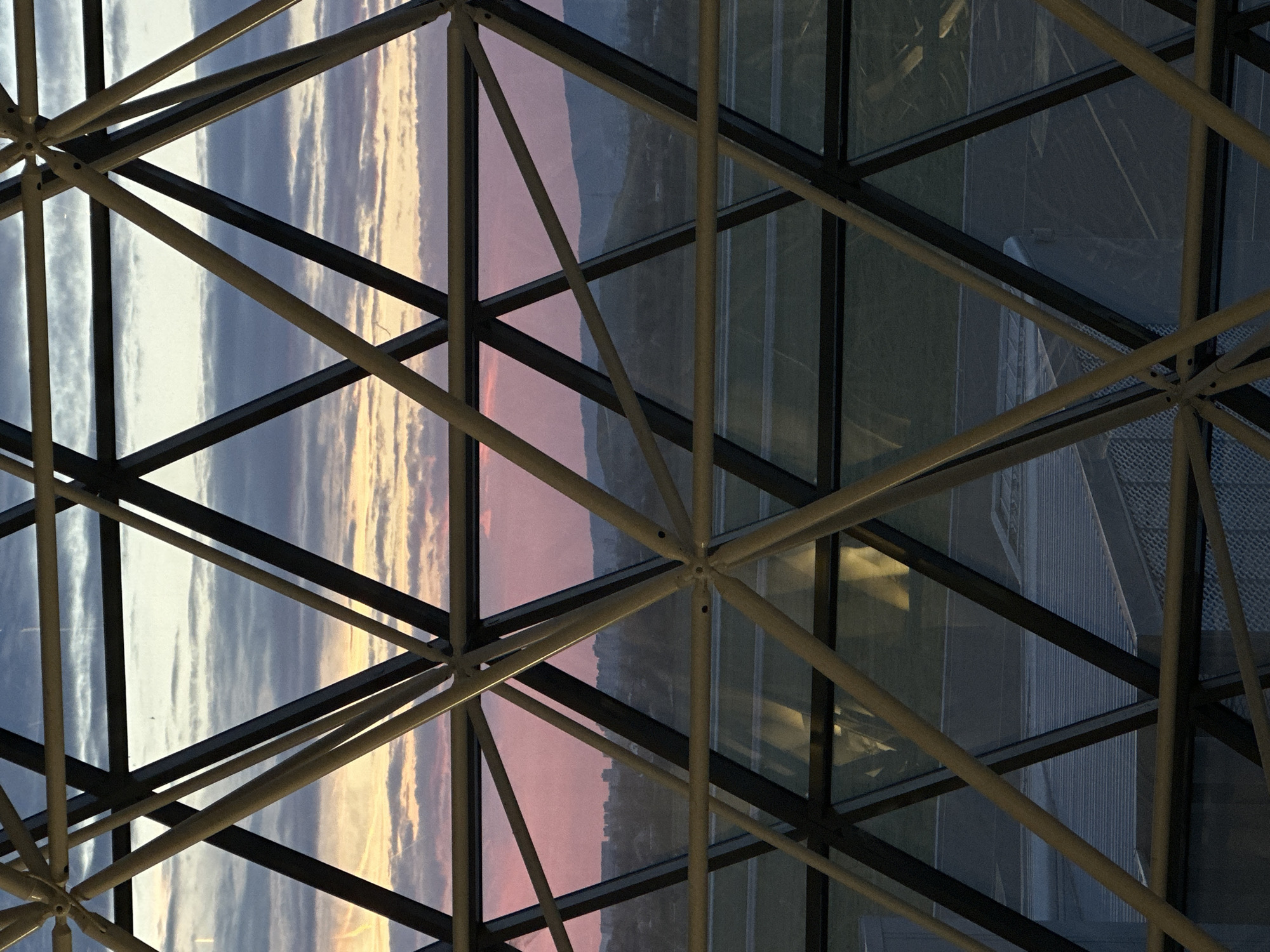 The image shows a close-up of a metal lattice structure, possibly part of a building's framework, with a view through the structure to a sunset or sunrise. The sky is visible through the grid, with hues of white, yellow, pink, and blue. On the right side, building interiors or adjacent structures are visible. The location is City of Velika Gorica, Croatia.