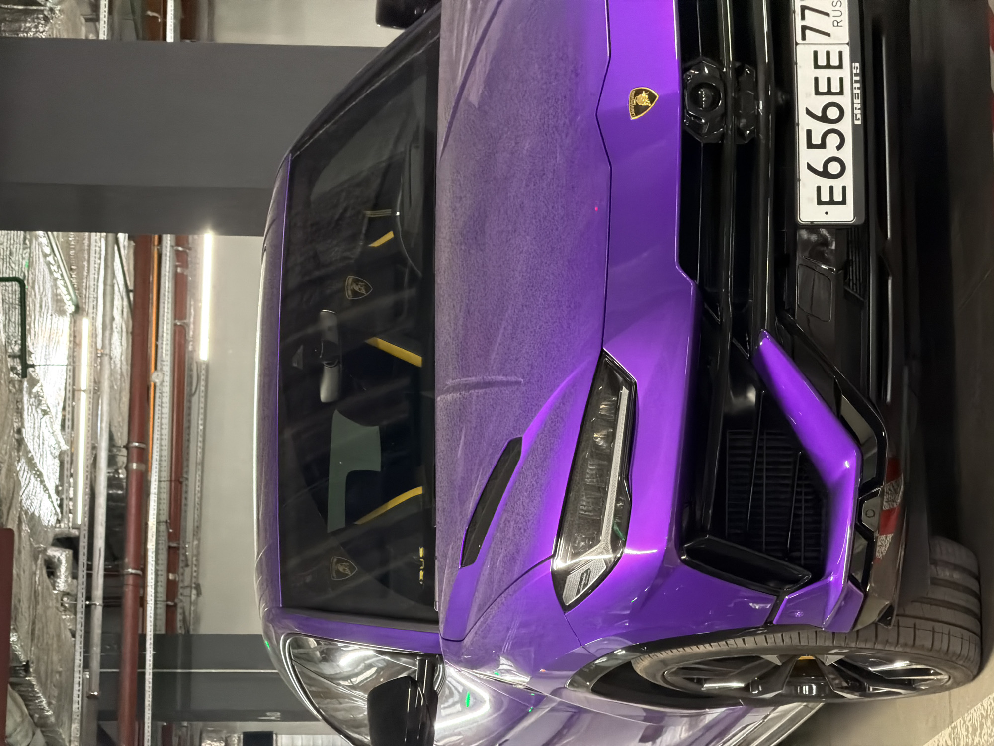 The image shows the front of a purple Lamborghini Urus parked indoors, likely in a garage or parking structure. The car has a license plate that reads "E656EE77" with "RUS" and "GREATS" printed on the plate, indicating it is registered in Russia. The environment appears to be utilitarian and functional, possibly an underground parking area in the city of Moscow.