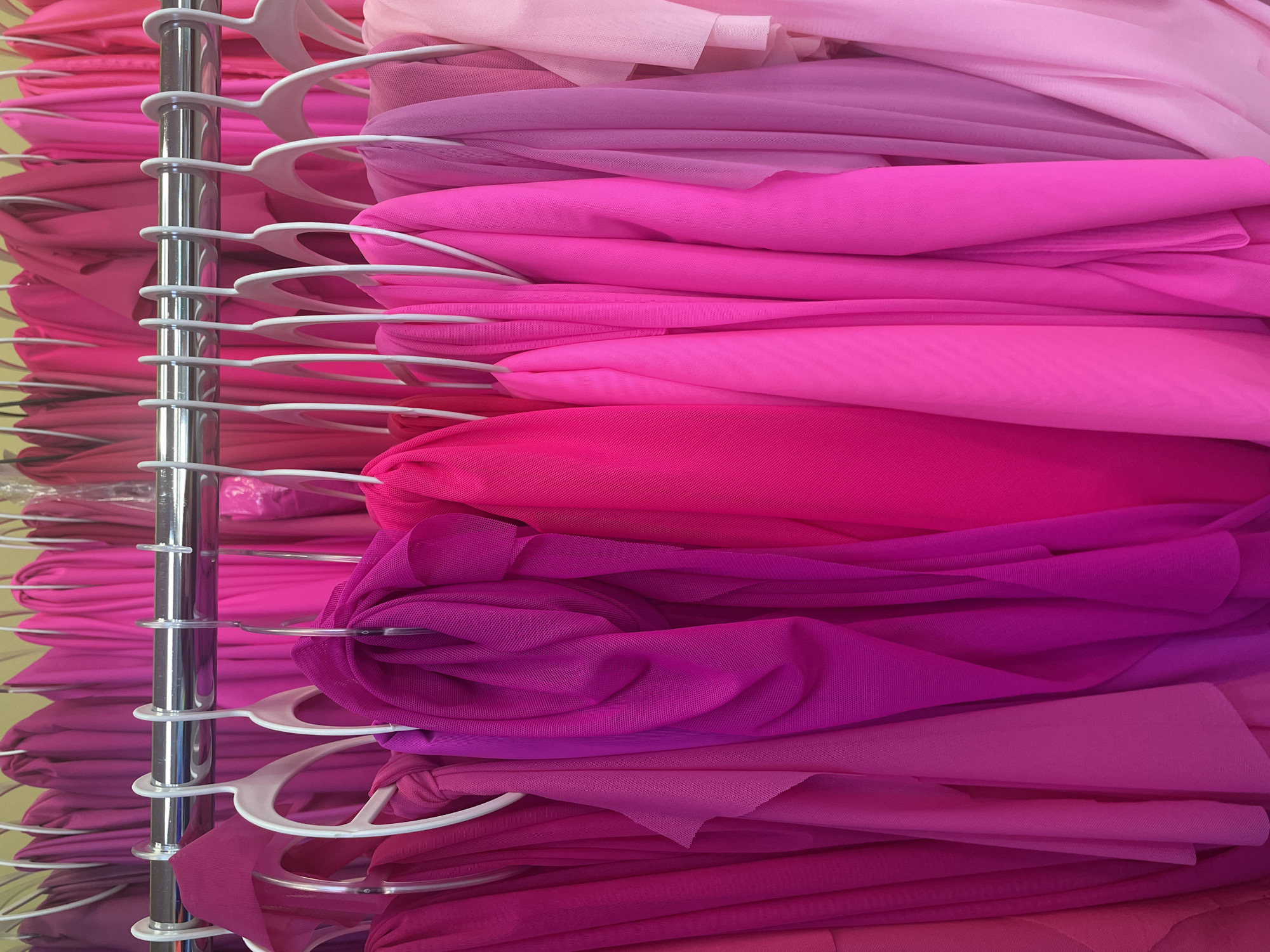 A close-up, eye-level shot shows a rack of sheer, flowing fabrics in various shades of pink. The fabrics are hung on white hangers from a silver metal rod. The fabrics are folded and draped, creating a textured appearance. The lighting is bright and even, highlighting the colors and textures of the material. There are no people or animals visible in the image. The background is out of focus but appears to be a wall or indoor setting.