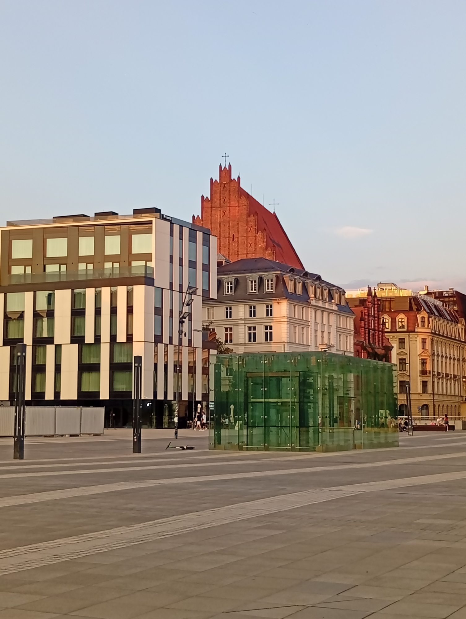The image depicts a modern plaza in Wrocław, Poland, with a contemporary building featuring a black and white facade and large windows in the foreground. To the right, a transparent glass structure with green tinted panels stands on the plaza. Behind these, older, historic buildings with red roofs and intricate architectural details are visible, including a tall brick building with a prominent red roof and a steeple topped with a cross. The sky is clear and pale blue, suggesting it is either early morning or late afternoon. The plaza floor is paved with grey and white rectangular tiles. In the middle ground, a black electric scooter lies on its side on the paving. Several indistinct figures are present, including two individuals walking in the background near a bench and a person in a wheelchair. The scene conveys a sense of urban space that blends modern and historical architectural styles.