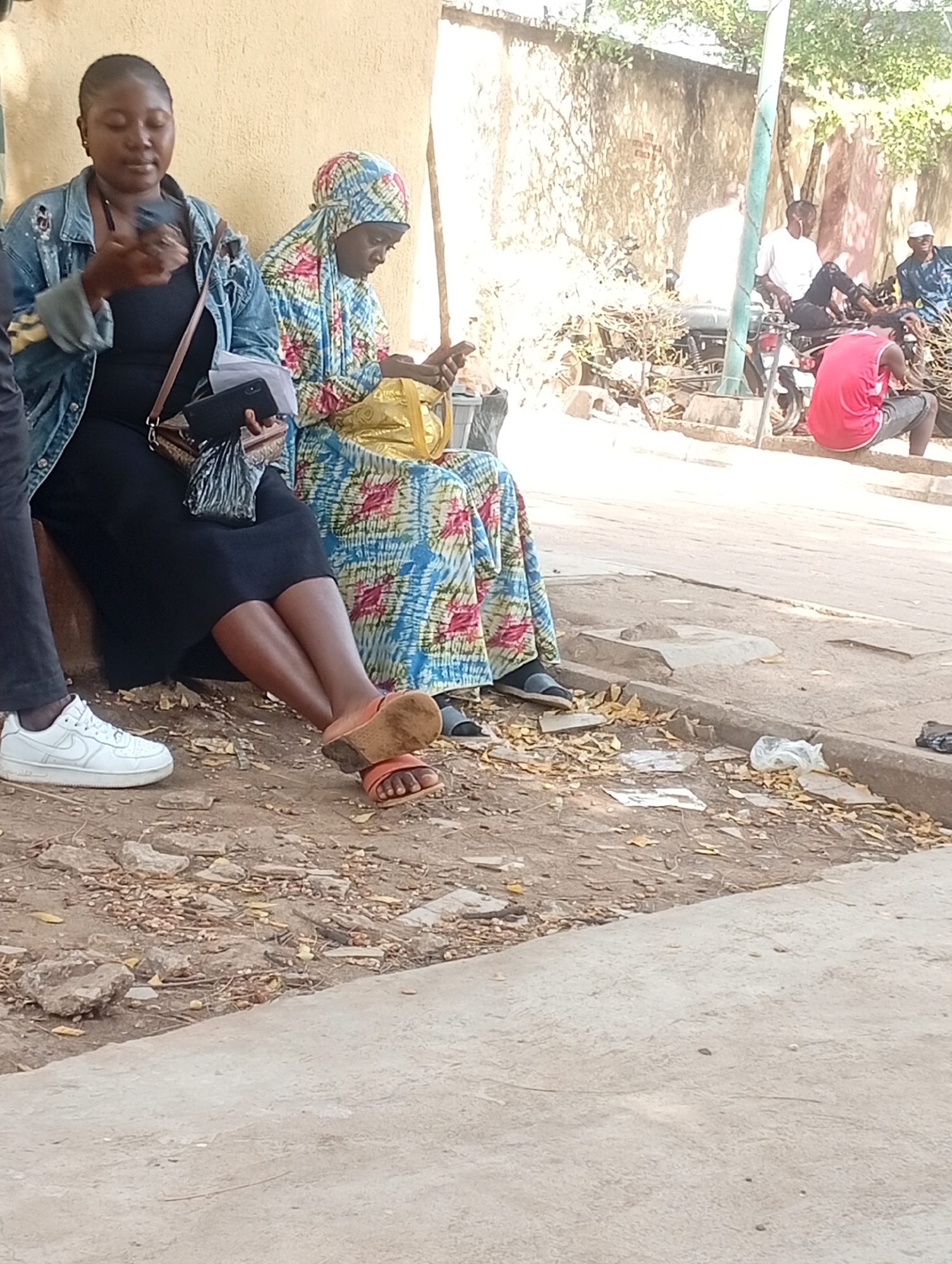 This image captures an outdoor scene in Barden Yana, Nigeria, featuring a group of people in a casual, dusty environment during daylight hours.
In the foreground, two women are seated. The woman on the left wears a denim jacket over a black top and skirt, with white Nike Air Force sneakers on her feet. She holds a dark smartphone in her left hand and a brown crossbody bag rests beside her. Her expression is neutral as she looks downwards. Next to her, another woman is dressed in a vibrant, multi-colored patterned fabric, including a head covering. She also looks down at a dark object, likely a phone, held in her hands. Her feet, clad in brown open-toed sandals, are visible on the dusty ground.
The ground surface is rough and unpaved, covered with dry leaves, small stones, and some scattered debris like plastic. A concrete curb separates this area from what appears to be a slightly raised, smoother concrete path or roadside on the right.
In the mid-ground and background, several men are visible. Some are seated on or near motorcycles, while others stand or sit on a low wall or curb. One man, seen from the back, wears a red shirt and is perched on a concrete ledge. The background includes a light-colored, somewhat weathered wall and sparse vegetation, suggesting a semi-urban or roadside setting. The overall activity appears to be one of waiting, resting, or socialising casually. The time of day is clearly daytime, indicated by the bright, even light and the distinct shadows. No specific emotional expressions are clearly discernible among the individuals; most appear calm or engrossed in their activities.