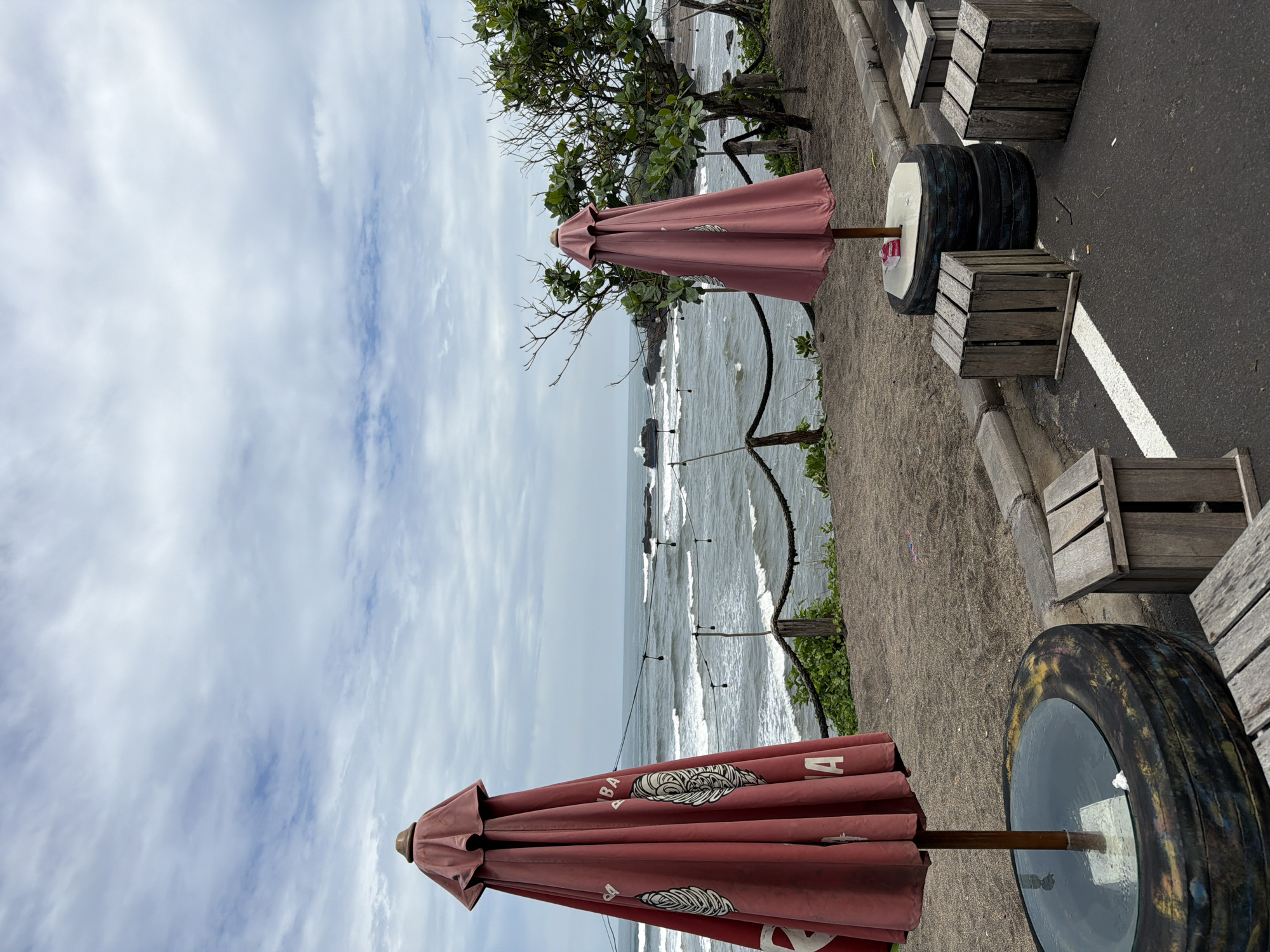 The scene depicts an outdoor seating area overlooking a coastline. There are two closed red umbrellas with decorative logos along the sides. Tables made from tires with glass tops are placed between wooden crate seating along a paved walkway. A tree is positioned between the umbrellas. A rope railing separates the walkway from the beach below. The ocean has visible waves. The sky is overcast with light and dark clouds. This scene is likely in or around Mengwi, Indonesia, given the provided context.