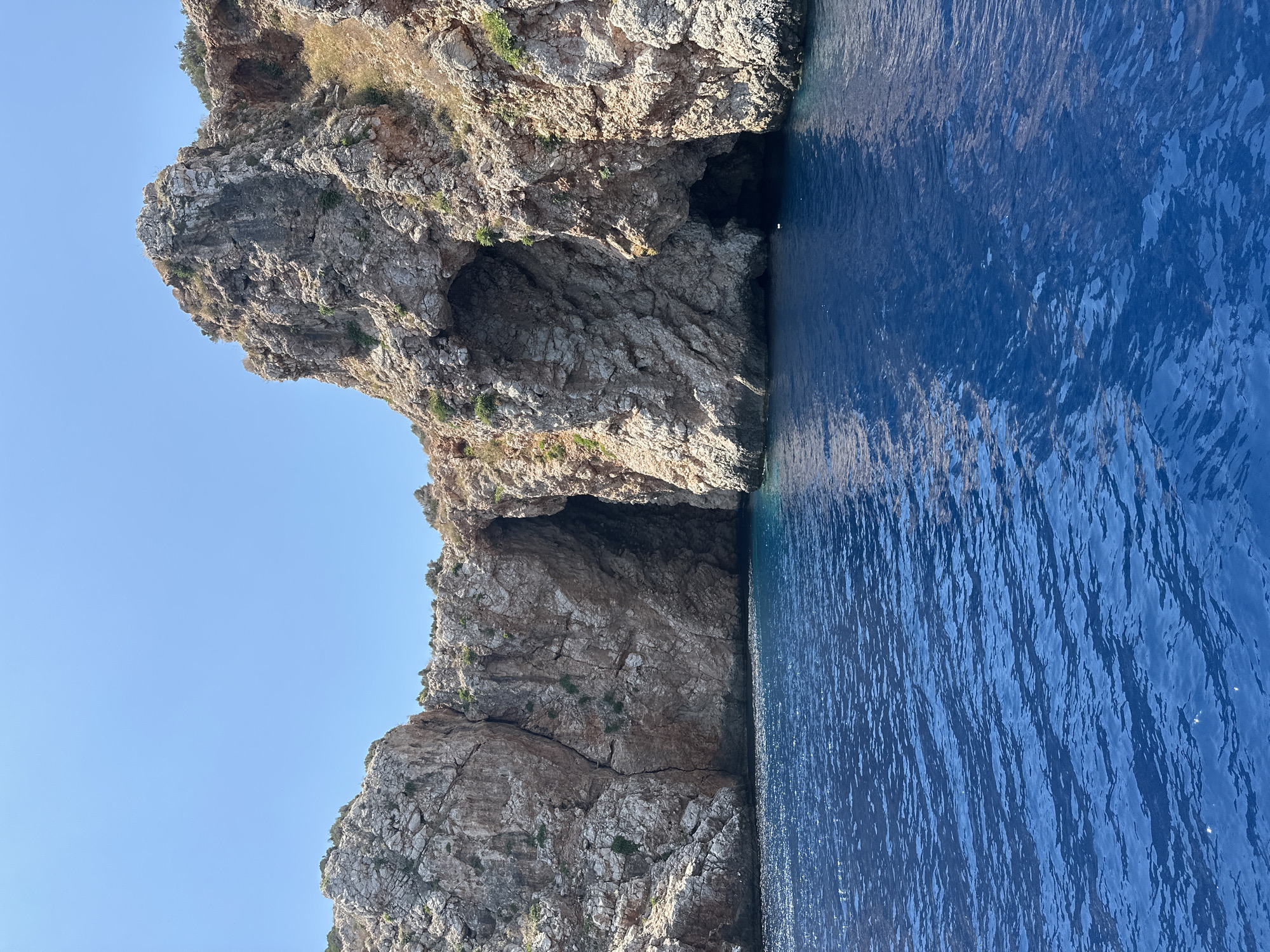 The image depicts a coastal scene featuring rugged, rocky cliffs with several cave openings carved into them. The sea in the foreground is a vibrant, deep blue, with gentle ripples on its surface. The cliffs are illuminated by bright sunlight, highlighting their rough texture and casting shadows within the cave entrances. Sparse vegetation, consisting of dry grasses and small bushes, grows on the rocky surfaces. The sky above is clear and a uniform light blue. The perspective suggests the photo was taken from a boat on the water, looking towards the cliffs and caves.