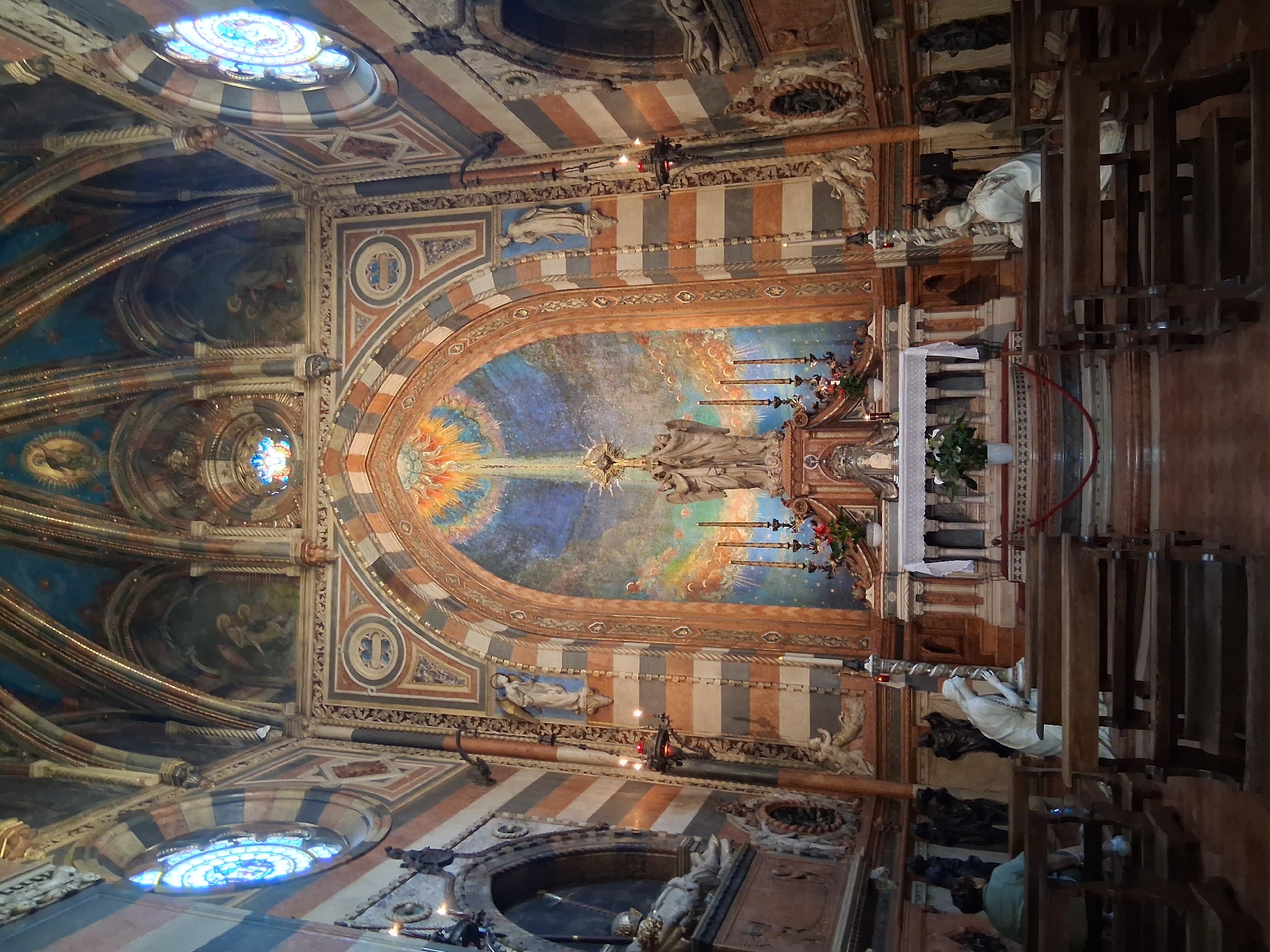 The image depicts the interior of the Scrovegni Chapel in Padua, Italy. The chapel is ornately decorated with frescoes and architectural details. In the foreground, several rows of wooden pews are visible, some occupied by individuals. To the right, a white statue of a kneeling figure is positioned near the pews. The central focus of the image is the altar area, which is adorned with a large fresco above it, depicting a celestial scene with figures and a beam of light. Statues of religious figures are present on either side of the altar. The walls and ceiling of the chapel are intricately painted with religious scenes and decorative patterns. Stained-glass windows with blue and white patterns are visible on the upper left and right sides of the image. The overall impression is one of solemnity and religious reverence.
