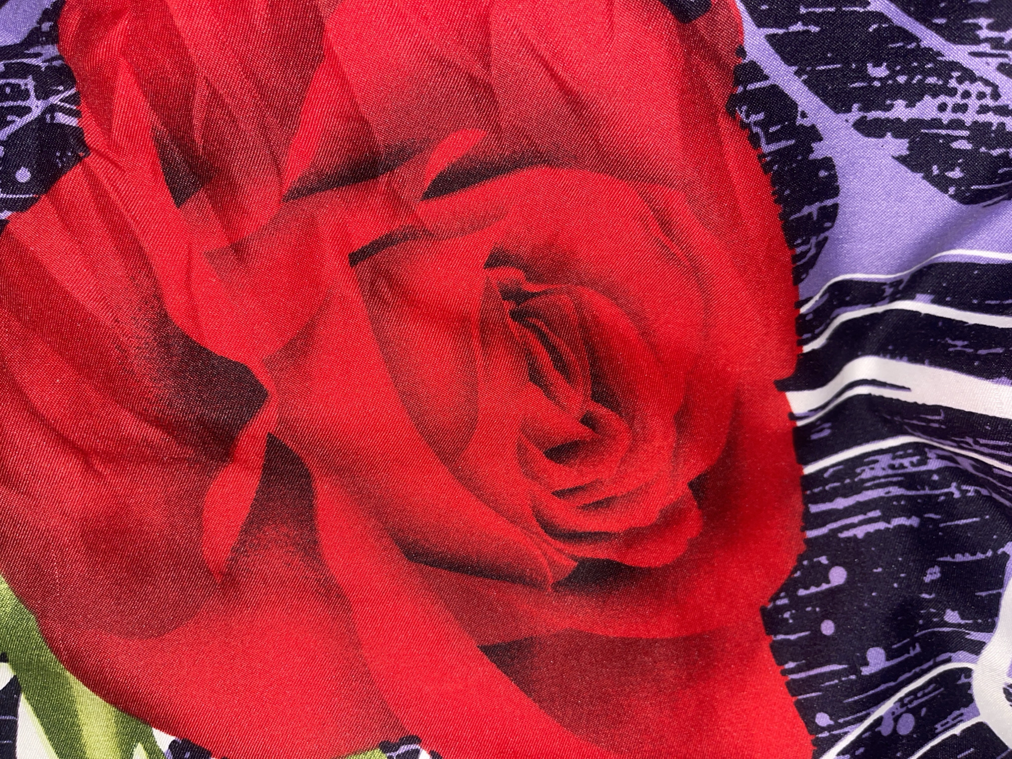 This is a close-up, abstract shot of fabric with a floral print. The dominant feature is a large, vibrant red rose with deep shadows and highlights that emphasize its texture and layers of petals. The rose is partially overlaid by another fabric pattern which consists of irregular black and purple splotches interspersed with thin white lines, resembling brushstrokes or abstract graffiti. In the lower portion of the image, there are also sections of a lavender-colored fabric with a similar abstract pattern. The lighting creates a slight sheen on the fabric, highlighting the weave. The overall impression is a textured, colorful, and somewhat abstract depiction of patterned textiles. There is no indication of any specific location within Minsk, Belarus, or any people or actions.