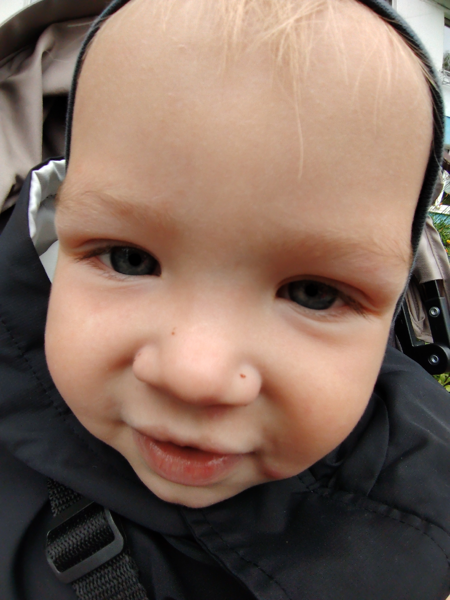 This is a close-up, wide-angle photograph of a young child's face, taken outdoors. The child, who appears to be an infant or toddler, is wearing a black jacket and a dark-colored hood that partially frames their face. The child's head is tilted upwards, and their gaze is directed towards the camera. Their eyes are open and appear to be blue or grey. The child has fair skin and light-colored hair, visible at the top of their head. The background is blurred, but suggests an outdoor environment, with hints of green foliage and possibly a stroller or baby carrier. The lighting is natural, indicating daylight. There is no information in the image to determine the specific location in Chaikovsky, Russia.