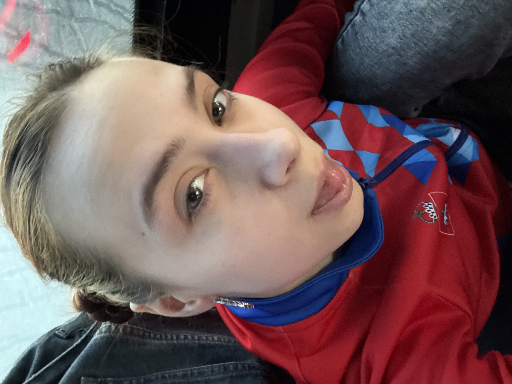 A close-up, slightly tilted headshot of a young woman. She has fair skin, light brown hair pulled back, and brown eyes. She is wearing a red jacket with blue trim and a blue and red geometric pattern on the upper left chest, along with a logo. She has a stud earring in her right ear. The background appears to be a mix of textures and colors, possibly indicating she is seated in a car or public transportation. Based on the information available, the location within the city of Berezniki, Russia, is unknown. Further information or a broader view of the image is needed to identify the location.