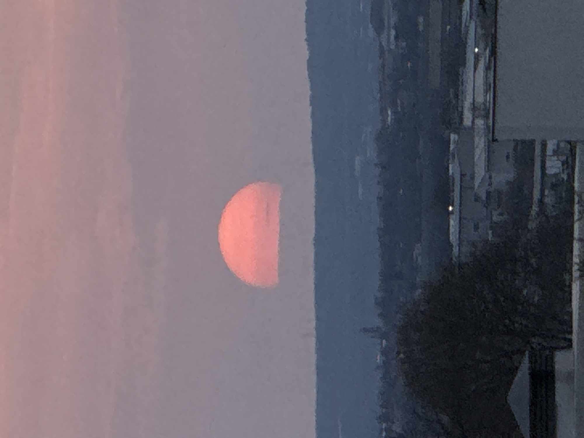 The image shows a half-obscured sun, orange-red in color, appearing on the left side of the frame. To the right of the sun, there is a dark, obscured landscape, possibly a cityscape. A partial building structure is visible on the right. The perspective of the image is rotated to the left. Based on the sun's color and position, this is likely a sunrise or sunset in Varna, Bulgaria.