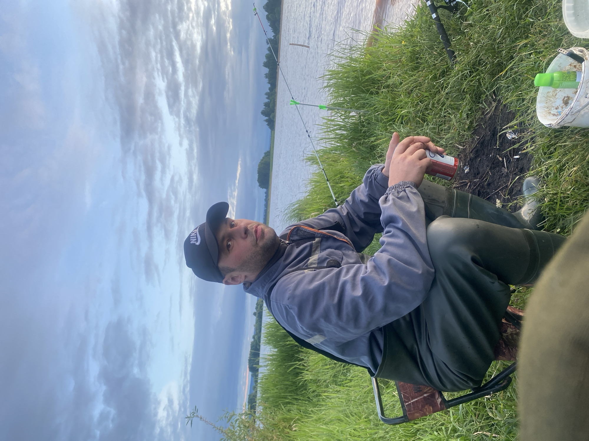 A male individual is seated on a portable camouflage-patterned folding chair on a grassy riverbank in Elovo, Russia. He wears a black Nike baseball cap, a grey work jacket with reflective accents, and dark green waders with integrated boots. He holds a red and white beverage can in both hands, resting on his lap, and looks directly towards the camera with a neutral expression. To his right, two fishing rods are propped up by green rod rests at the water's edge, extending into the wide body of water. The river has a slightly textured surface, and a distant treeline is visible on the opposite bank. The foreground shows tall green grass, a small area of disturbed earth with minor refuse, and a white bucket containing a green spray bottle and other items. The sky above is largely overcast with soft grey and blue clouds, indicating an outdoor setting during twilight hours.