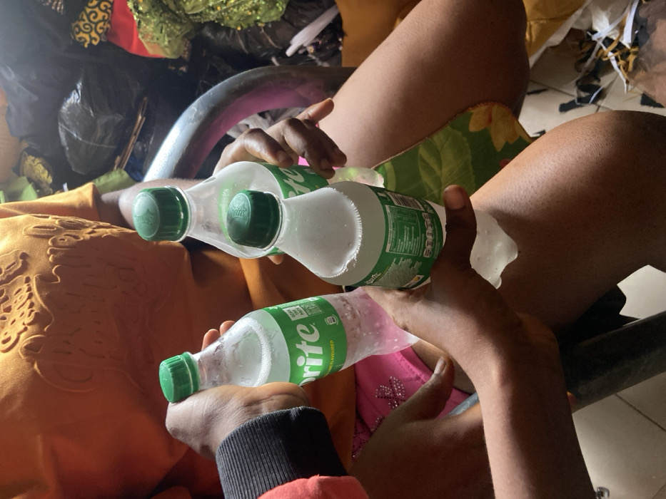 Two people holding Sprite bottles are seated next to each other. The bottles appear to be cold, with condensation forming on the outside. One person is wearing a gold-colored shirt or dress, while the other has on a black and red sleeve. The people are seated in metal chairs, with the legs of the person on the right visible. In the background, there is what appears to be clothing and other items.