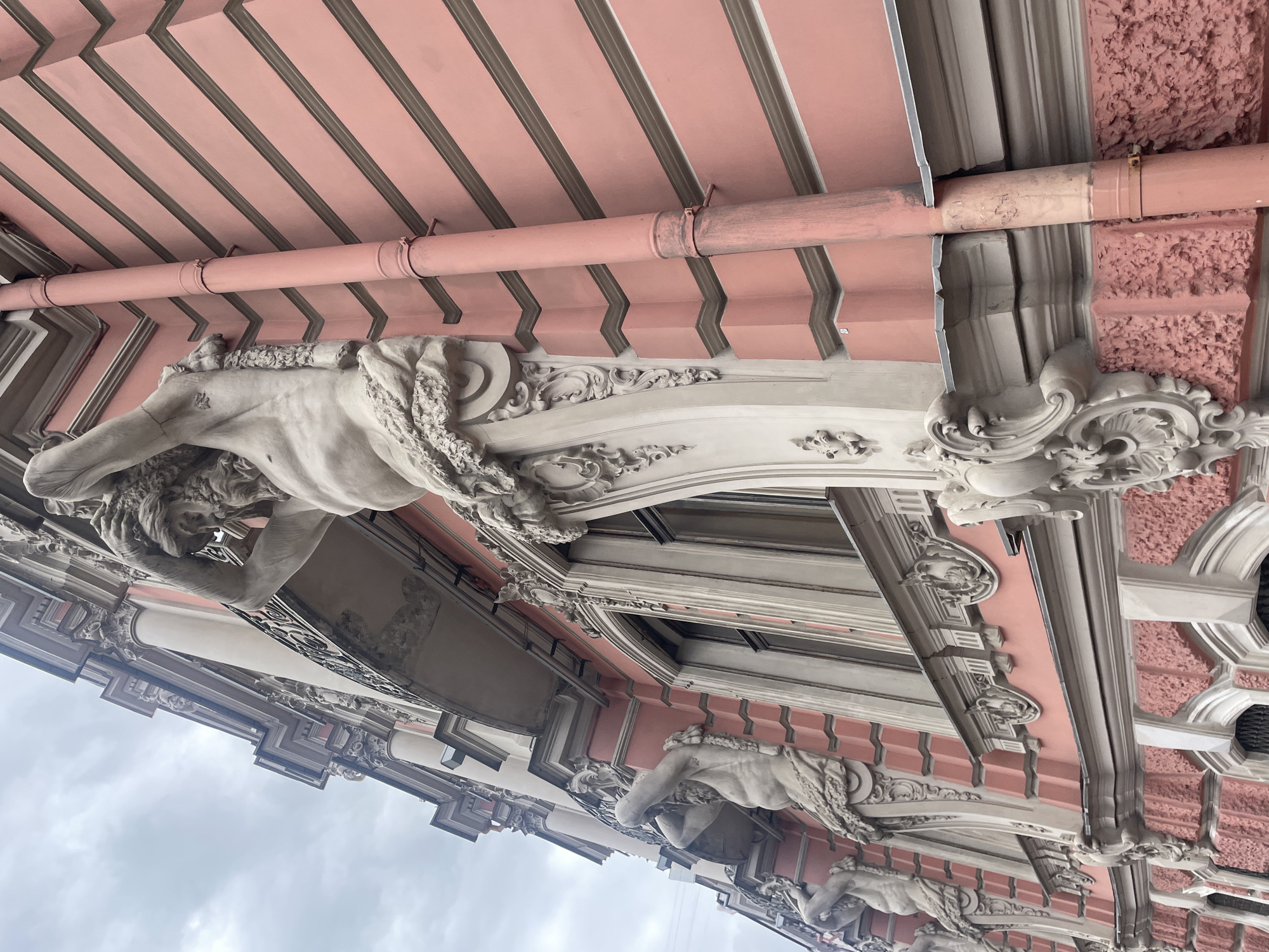 The image shows the exterior of the Singer House, also known as the Dom Knigi, located on Nevsky Prospekt in Saint Petersburg, Russia. The photograph focuses on the upper floors of the building, highlighting its ornate architectural details. Specifically, it features several Atlantes figures, which are muscular male sculptures, supporting the structure above. These figures are carved in stone and are intricately detailed, with beards and powerful physiques. They are positioned in front of windows and balconies, contributing to the building's grand Baroque or Neoclassical style. The building's facade is painted a pale pink color, with contrasting grey decorative elements and horizontal banding. A rusty-colored downspout runs vertically along the side of the building, adjacent to one of the Atlantes. The sky above is overcast with grey clouds. The perspective is looking upwards, emphasizing the scale and grandeur of the architecture.
