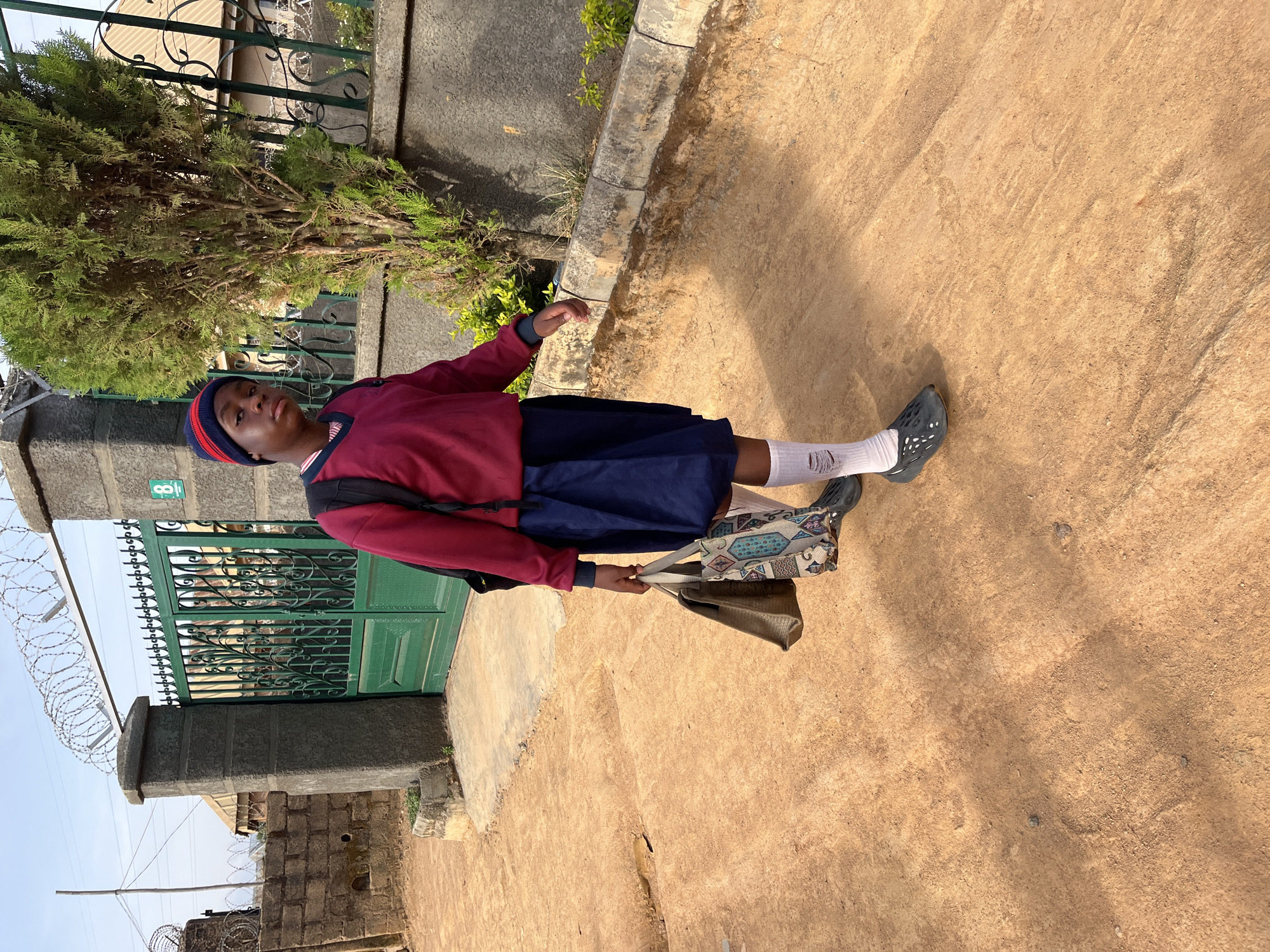 A young person, dressed in a school uniform consisting of a red sweater, navy blue skirt, and white socks, is standing on a dirt road next to a green gate with barbed wire in Jos, Nigeria. The person is carrying two bags, one patterned and one olive green. The background includes a tree, a concrete wall, and a metal fence. The gate is adorned with decorative metalwork and has a sign reading "8". The area appears to be a residential or institutional setting. The weather seems sunny, casting shadows on the ground.