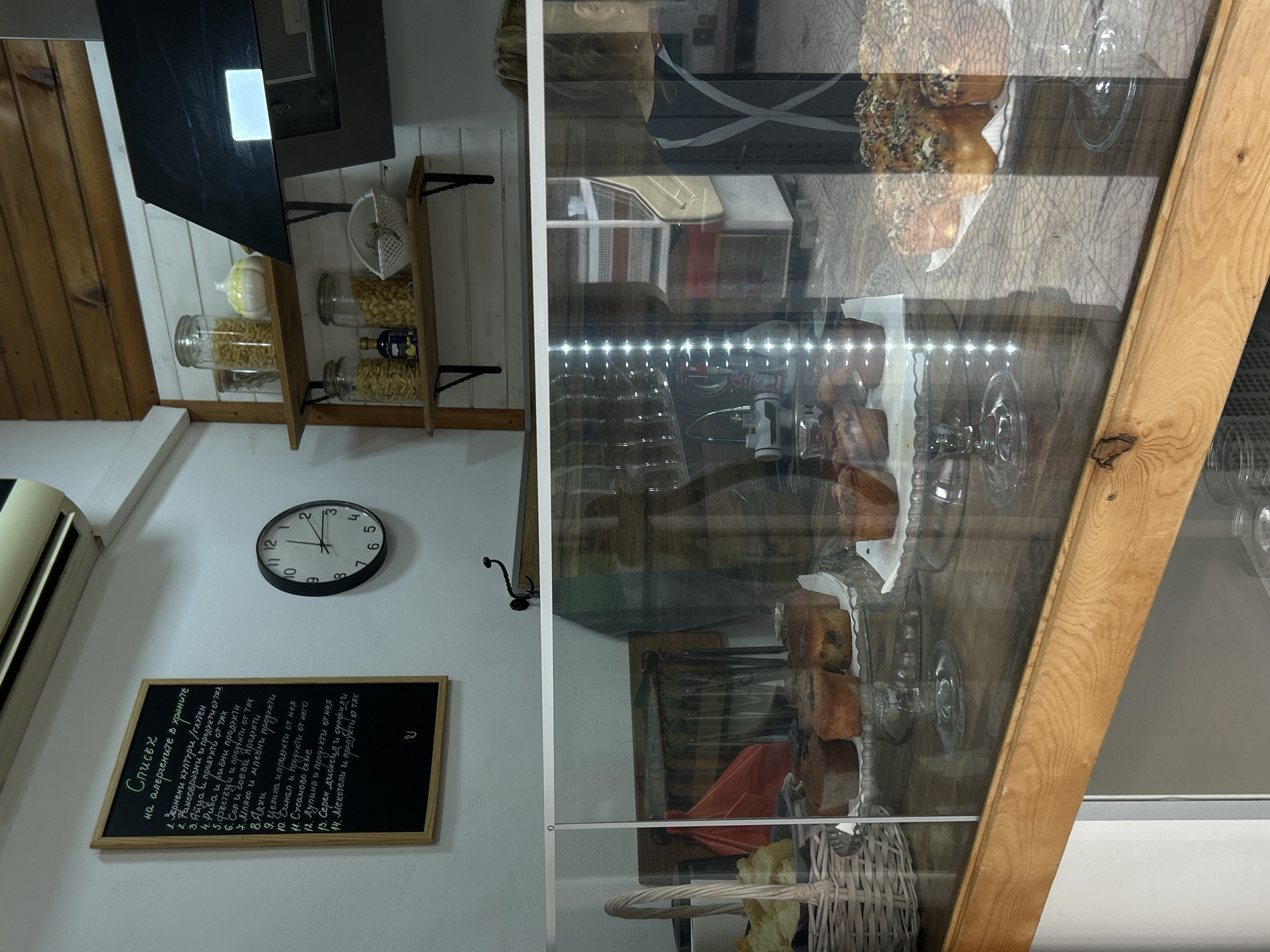 This appears to be an interior shot, likely of a small shop or bakery. The scene features a display case with various baked goods on multiple tiers. Items like pastries, bread, and possibly cakes are visible inside the display case. The display case is framed with light-colored wood and has a clear glass front.
On the left side of the image, attached to the wall, there is a clock with a black frame and white face. It shows the time as approximately 11:26. Below the clock, there is a chalkboard with a list written on it in Cyrillic script. Above the clock, shelves hold jars filled with pasta and other food items. A black range hood or kitchen appliance is mounted above the shelves.
The lighting in the image is a mix of natural and artificial light, with bright light emanating from the display case and softer light in the surrounding area. The wall is painted white and is likely located in Sofia, Bulgaria because the list on the chalkboard is written in Cyrillic.