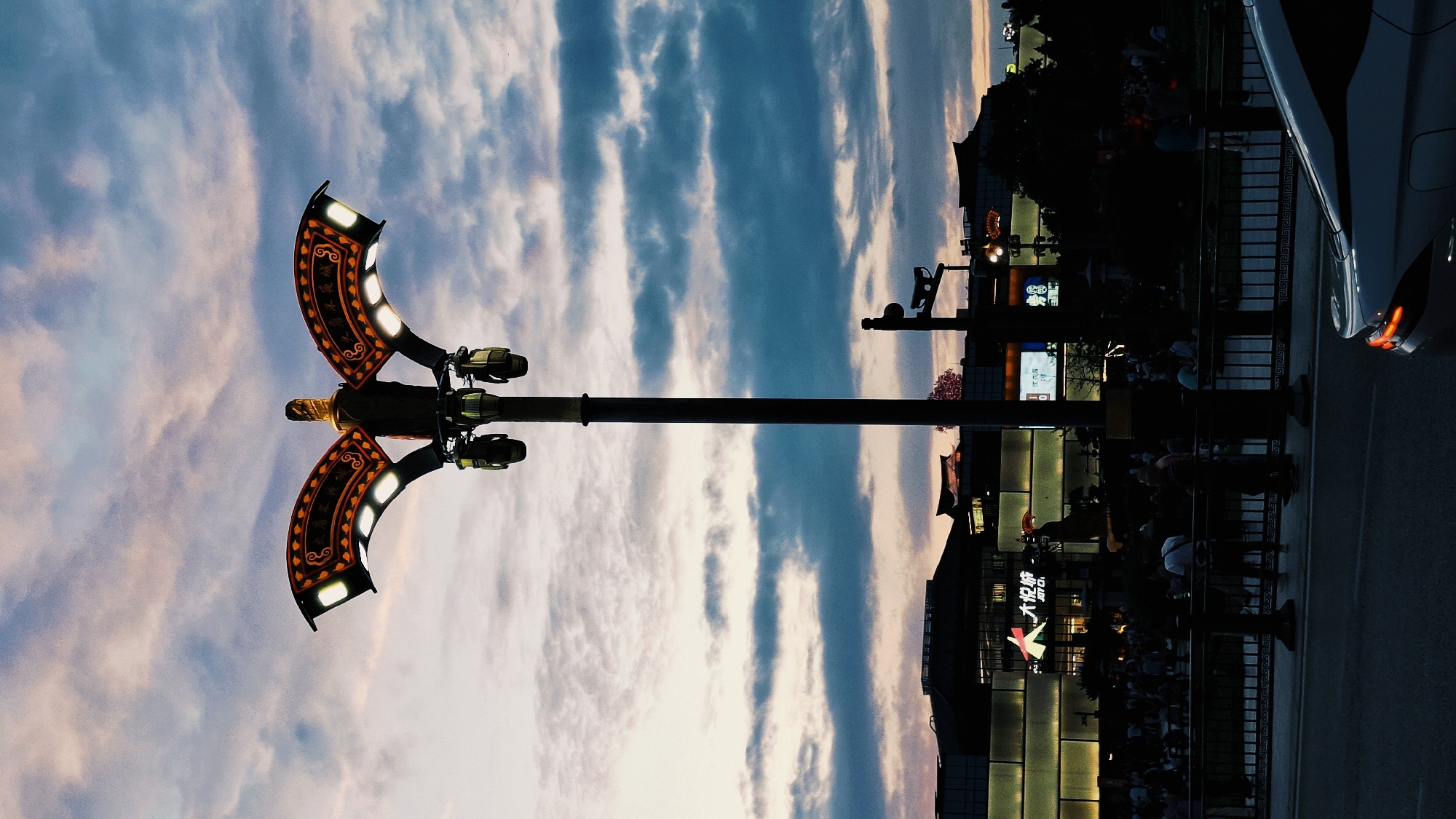 The image captures a street scene in Yanta District, China, oriented vertically. A decorative lamppost with two ornate, crescent-shaped light fixtures dominates the left side, silhouetted against a cloudy sky with hints of pink and blue. The light fixtures feature a pattern of orange and black.

On the right, a dimly lit street shows signs of activity. Buildings with lighted signs are visible, and a crowd of people stand behind a black metal railing separating them from a road. A portion of a car is visible on the right edge.