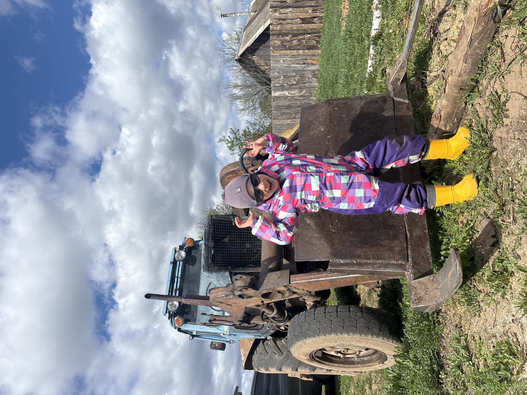 A young child wearing a purple, pink, and blue plaid snowsuit, a gray beanie, and yellow boots lies on top of a tractor in an outdoor setting in Ваньки, Russia. The child is smiling and holding up two fingers on each hand. The tractor is blue and brown with a black tire visible. Behind the tractor, there is a wooden structure, possibly a barn or shed, surrounded by grass and dirt. The sky is visible in the background, with a mix of blue sky and white clouds. The photo appears to be taken during the day.