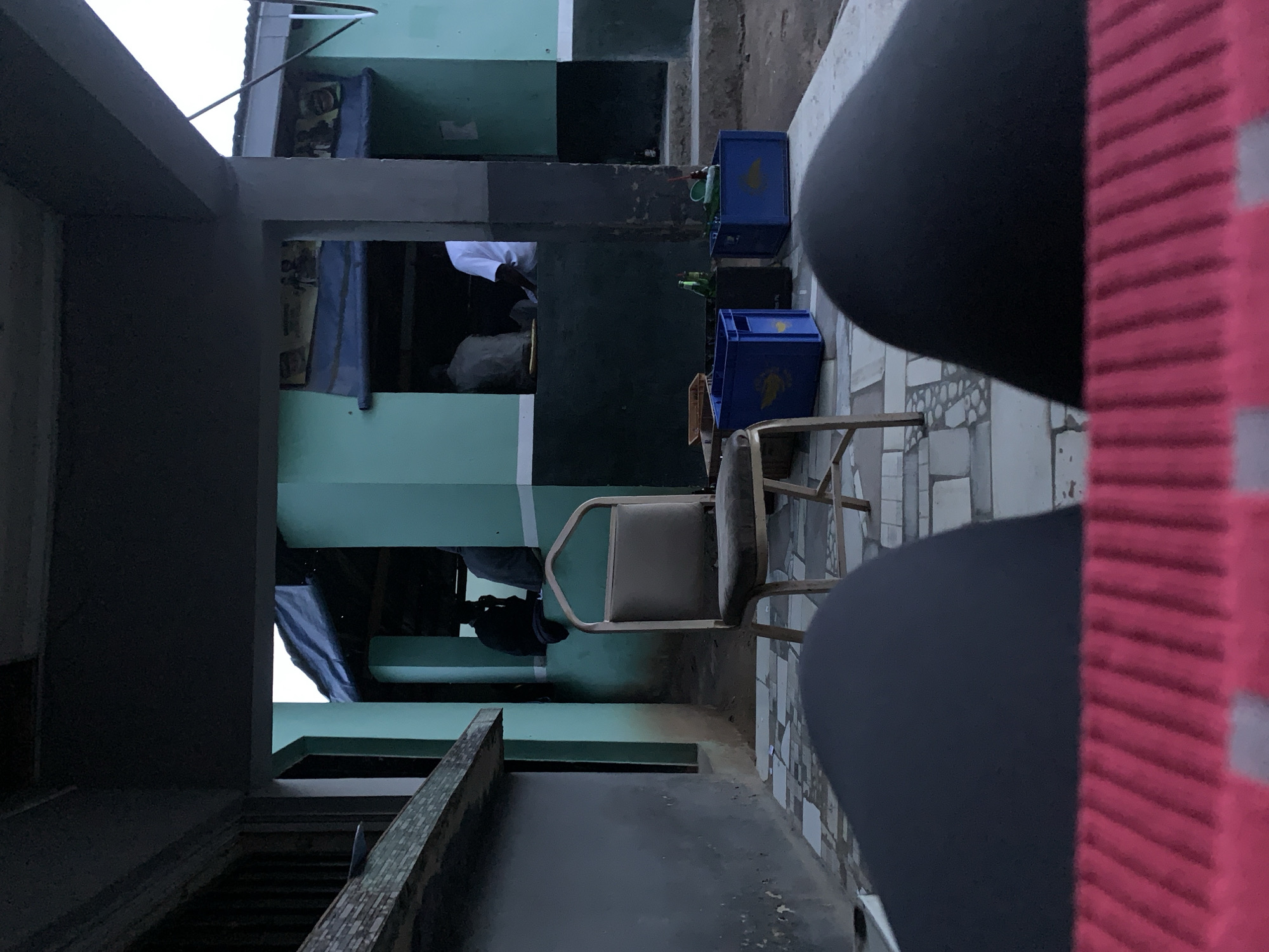 This image, captured in New Stadium, Nigeria, presents a low-angle view from a seated perspective, likely on a patio or outdoor bar area. In the immediate foreground, the bottom edge is blurred, showing a red, horizontally ribbed object and two dark, indistinct shapes that appear to be legs covered in dark fabric.
The midground features a patterned tiled floor, which is partially discolored in areas. An unoccupied, light-brown upholstered chair with a gold-colored metal frame stands slightly to the left of the center. To the right of the chair, three crates are visible on the ground: two blue plastic crates with handles, each containing multiple green bottles (likely beverages), and one dark-colored crate partially obscured between the two blue ones. A small, light-colored bowl or dish rests next to the leftmost blue crate.
The background reveals a series of buildings, possibly a commercial establishment or communal area, painted in a distinctive two-tone scheme of light teal/mint green on the upper sections and dark grey/black on the lower sections, separated by a white horizontal stripe. Several open windows or doorways expose the interior, where multiple individuals are seated or standing. One person on the left is mostly silhouetted in dark clothing. Another individual in the middle is seen from behind, wearing a white shirt. Faded posters or signs, depicting figures, are visible above the openings. The rooflines of the buildings are dark against a lighter, possibly overcast, sky. A white vertical pipe or conduit runs along the exterior wall on the far right. The ground beyond the tiled area appears to be uneven dirt or concrete. The overall lighting is subdued, suggesting either an overcast day or the approach of dusk.