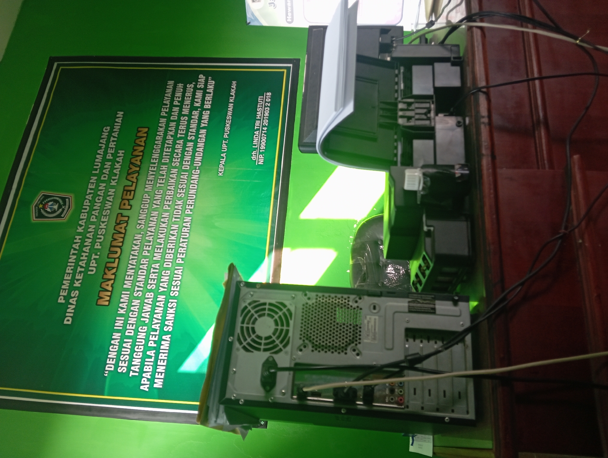 In the city of Tegalciut, Indonesia, a dimly lit indoor scene captures a desk with computer equipment and a large green poster. On the left, the rear of a beige computer tower is visible, displaying cooling fans and various ports. To its right, a printer with its lid open shows internal components and ink cartridges. Cables snake across the desk. Behind the equipment, a prominent green poster with white and yellow text is mounted on a green wall, featuring text that appears to be an official notice or announcement in Indonesian, including a logo at the top. The lighting is uneven, with shadows and highlights suggesting an artificial light source. The overall atmosphere is functional and appears to be an office or administrative space.