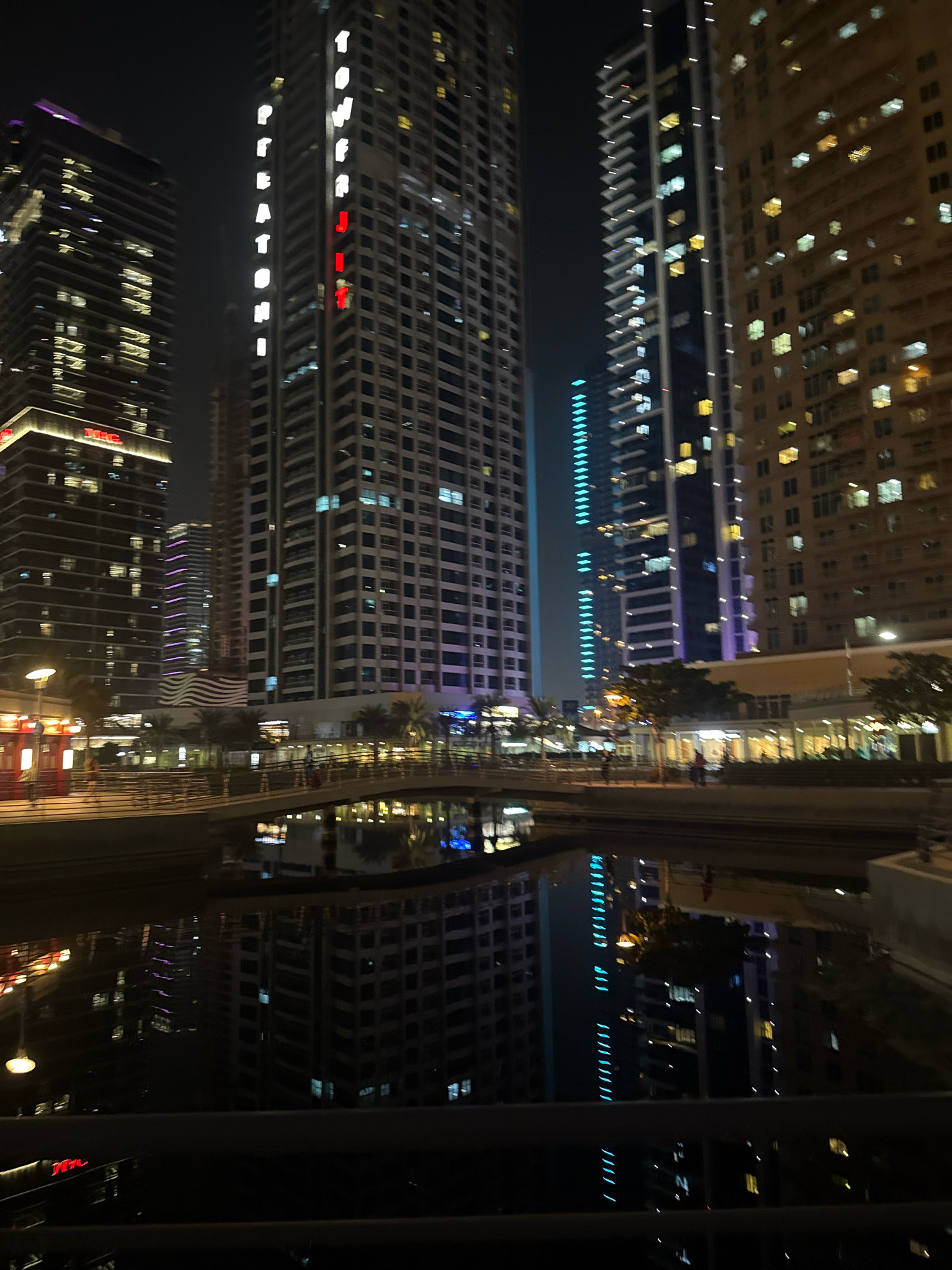 This is a nighttime, medium-shot, eye-level photograph of a canal in Dubai, United Arab Emirates, with a focus on the surrounding skyscrapers and their reflections in the water. The image is characterized by a dark sky and the bright, varied illumination from the buildings.
In the background, several tall, modern skyscrapers dominate the scene. On the left, a dark-colored building with numerous illuminated windows stands prominently. To its right, a taller, lighter-colored skyscraper features vertical lines of white and red lights that spell out words, with "TOWER" and "RESIDENCES" partially visible. Further to the right, another very tall skyscraper displays rows of bright white lights, creating a striking vertical pattern. The building on the far right has a more textured facade, with many windows lit in warm yellow and orange tones. Other, less distinct buildings are visible in the mid-ground and background, some with purple and blue lighting.
In the foreground, a dark canal runs horizontally across the image. The water is still, creating clear reflections of the buildings and the bridge above. A pedestrian bridge, illuminated by warm lights, spans the canal. Several indistinct figures can be seen walking on the bridge. Along the banks of the canal, there are low-rise structures with bright lighting, possibly shops or restaurants, and palm trees adding a touch of greenery. The overall atmosphere is one of urban nightlife, with the reflections in the water adding depth and visual interest.