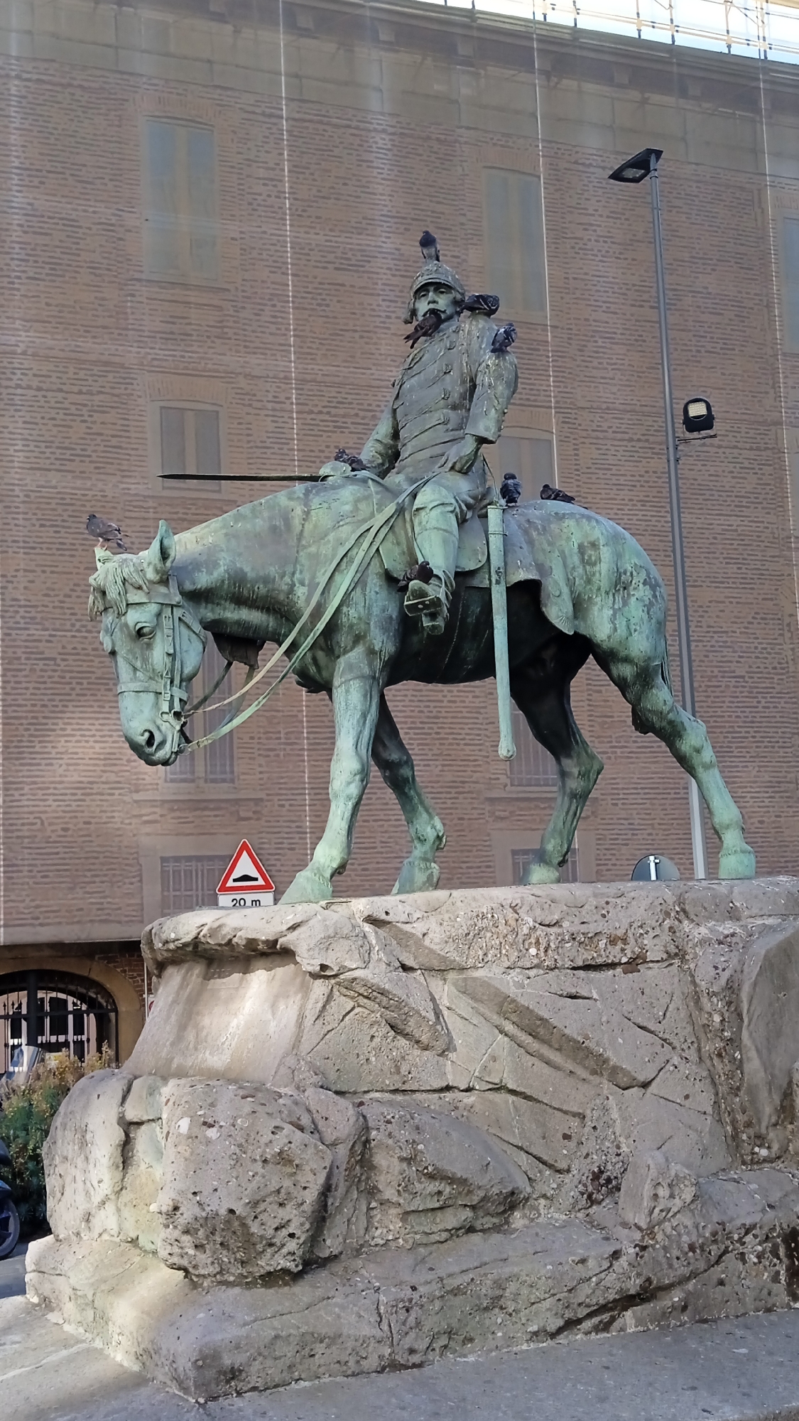 The image shows a bronze equestrian statue of a man in military attire, possibly a general or a king, adorned with pigeons. The statue is mounted on a large stone pedestal. The background features a brick building with arched windows covered by scaffolding and a streetlamp. A traffic sign indicating a speed bump at 20 meters is visible on the pedestal. The overall lighting suggests it is daytime. This monument is located in Piazza del Duomo, Milan, Italy, and is dedicated to King Umberto I of Italy.