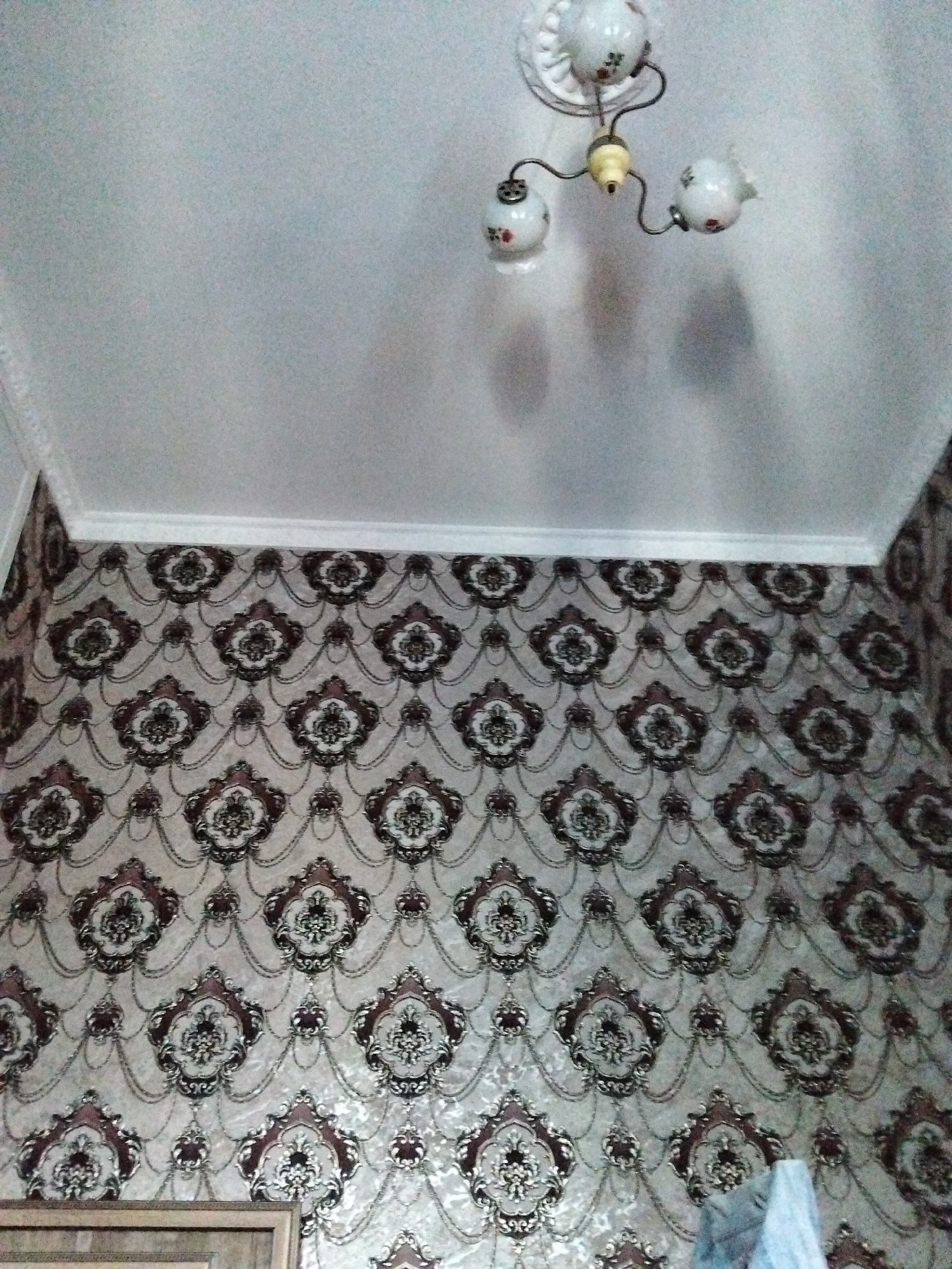 The image shows a close-up view of a wall with ornate wallpaper and a ceiling-mounted light fixture. The wallpaper features a repeating pattern of large, dark-bordered floral medallions interspersed with smaller decorative elements and chain-like motifs against a lighter background. The ceiling is white and has a decorative molding where it meets the wall. A vintage-style light fixture with three porcelain shades, each adorned with a floral design, is attached to the ceiling. The light fixture has an off-white base and metal arms. The scene appears to be indoors, likely in a residential setting. The image is taken from a low angle, looking upwards. The overall impression is one of a decorated interior space.