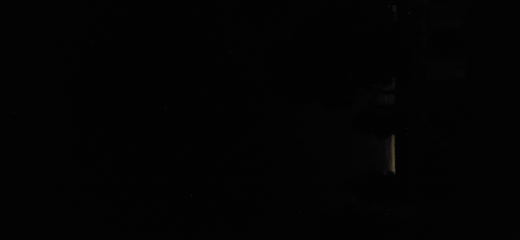 The image is a low-light, black and white photograph of a street scene in Eeklo, Belgium. The street is empty, and the only visible light source appears to be a faint, yellowish glow emanating from a horizontal element in the lower portion of the frame, likely a street light or illuminated signage. The background is predominantly dark, suggesting it is nighttime or the area is poorly lit. The overall impression is one of quiet and stillness.