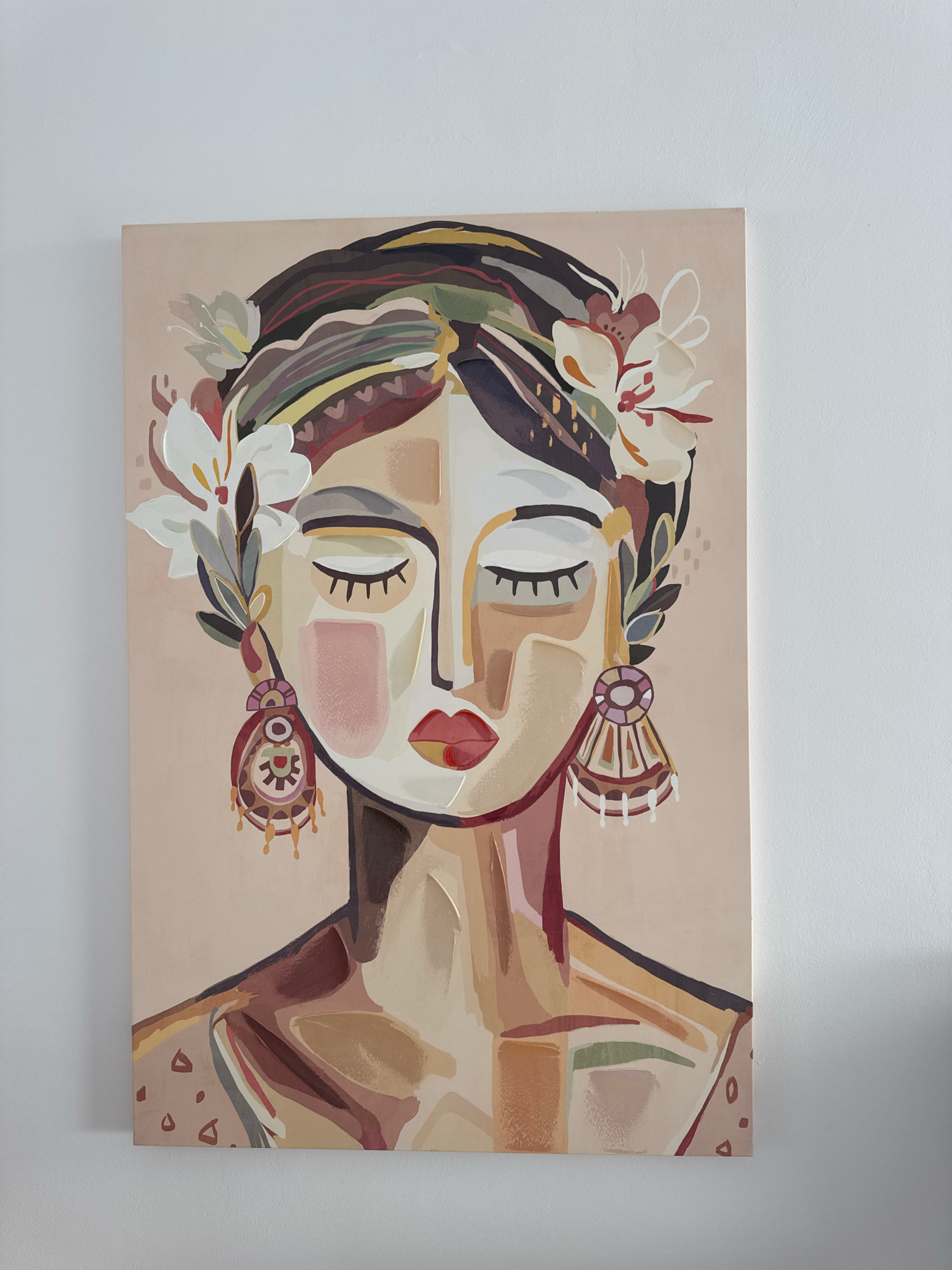 A framed canvas painting of a woman's face is mounted on a white wall. The painting features a stylized portrait with closed eyes, adorned with flowers and large, decorative earrings. The colors are muted and earthy, with a dominant beige background. The style is abstract and illustrative. The painting is illuminated by natural light, casting a subtle shadow on the wall to its right. The image is captured from a straight-on perspective, slightly angled from above, showing the top edge of the canvas and its mounting on a wall in Limassol, Cyprus.