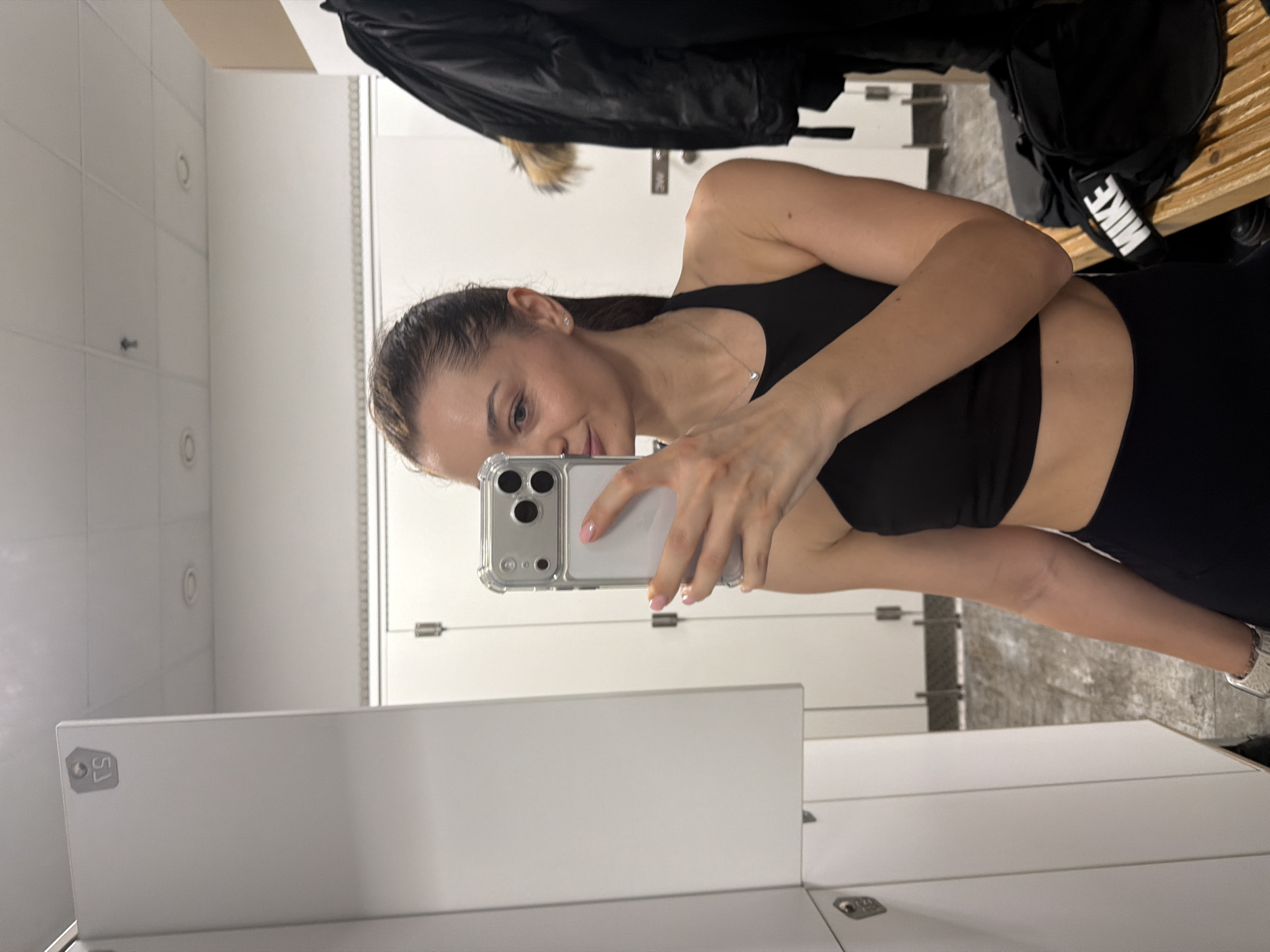 A woman is taking a selfie in what appears to be a locker room. She is wearing a black sports bra and leggings. She is holding a silver-colored iPhone in her left hand. The background shows white lockers. A black jacket and a black Nike bag are visible on the bench. It is impossible to determine the location in the city of Varna, Bulgaria from the image alone.