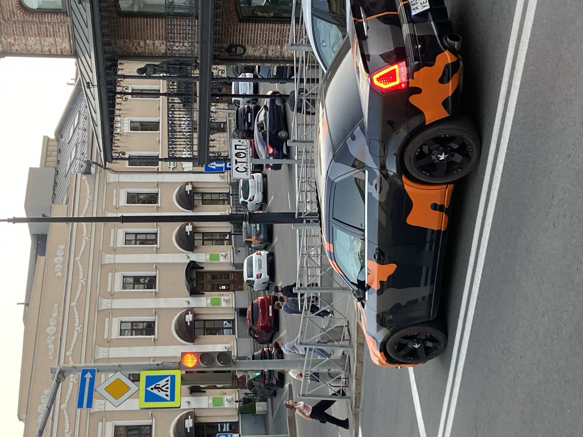 The image shows a street scene in Kazan, Russia, oriented vertically. The street is lined with buildings, including a beige building with ornate detailing and a darker, metal-clad structure with a balcony. Several cars are parked along the street, including a black car with orange camouflage accents. A pedestrian crossing sign and a traffic light are visible. People are walking along the sidewalk. A "СТОП" (Stop) sign is visible in Russian. Other parked cars are visible further down the street.