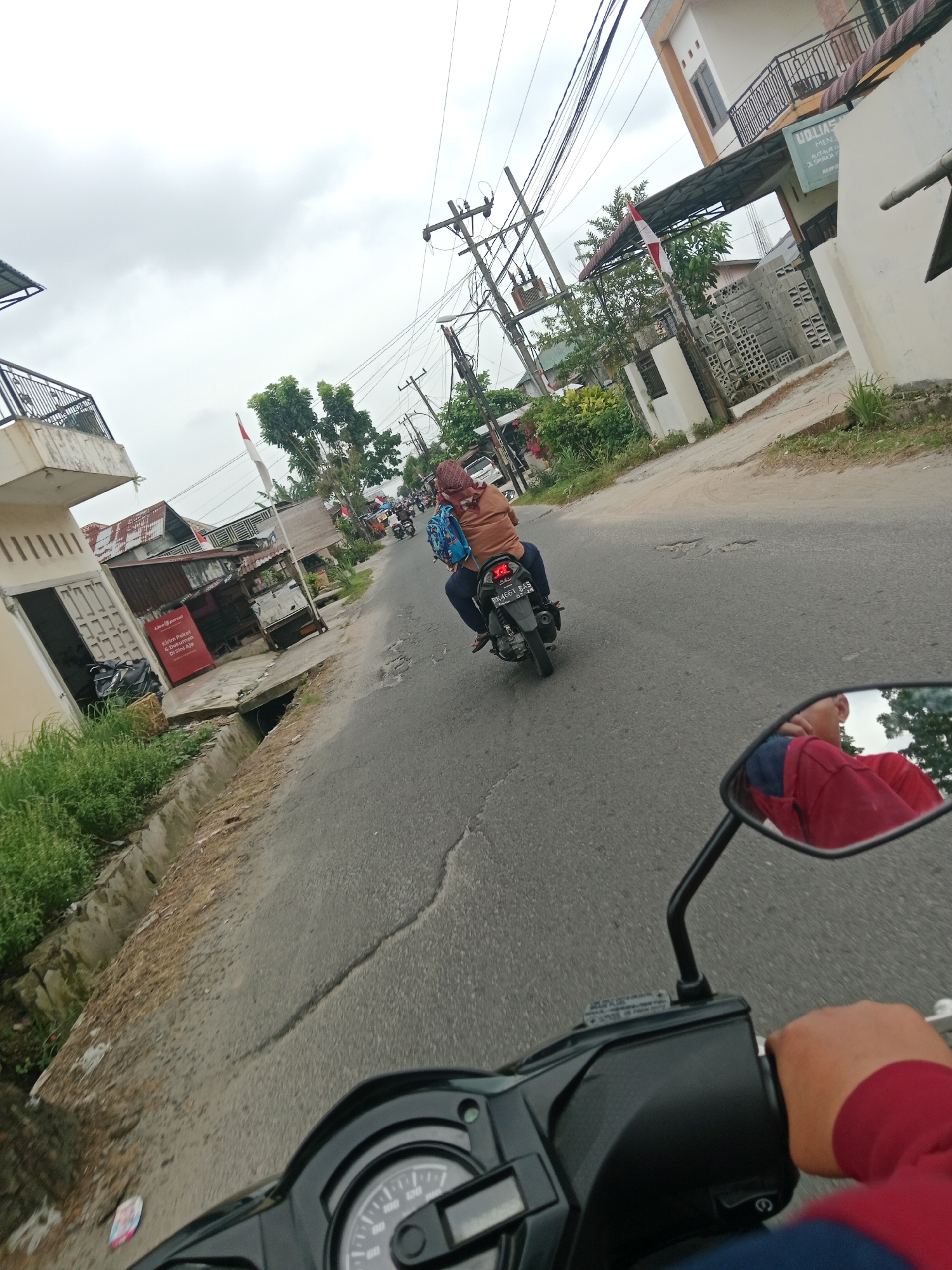 The image shows a view from the perspective of someone riding a motorcycle on a street in the City of Binjai, Indonesia. In the foreground, the handlebars and a portion of the motorcycle's dashboard are visible, along with the rider's hand in a red jacket. A side mirror reflects the rider's head, also wearing a red garment. Ahead on the road, another motorcycle is moving away, carrying two people. The person in front is wearing an orange top and a headscarf, with a blue backpack. The road is paved and shows signs of wear and tear, with a concrete drainage ditch along the side. On the left side of the road, there are several buildings, including a house with an open doorway and a small shop with a red sign that says "Kirim Paket & Dokumen Di Sini Aja". There are also trees, bushes, and power lines overhead. On the right side of the road, there is a building with a balcony and a sign that reads "UOLIAST MEN". The sky is overcast.