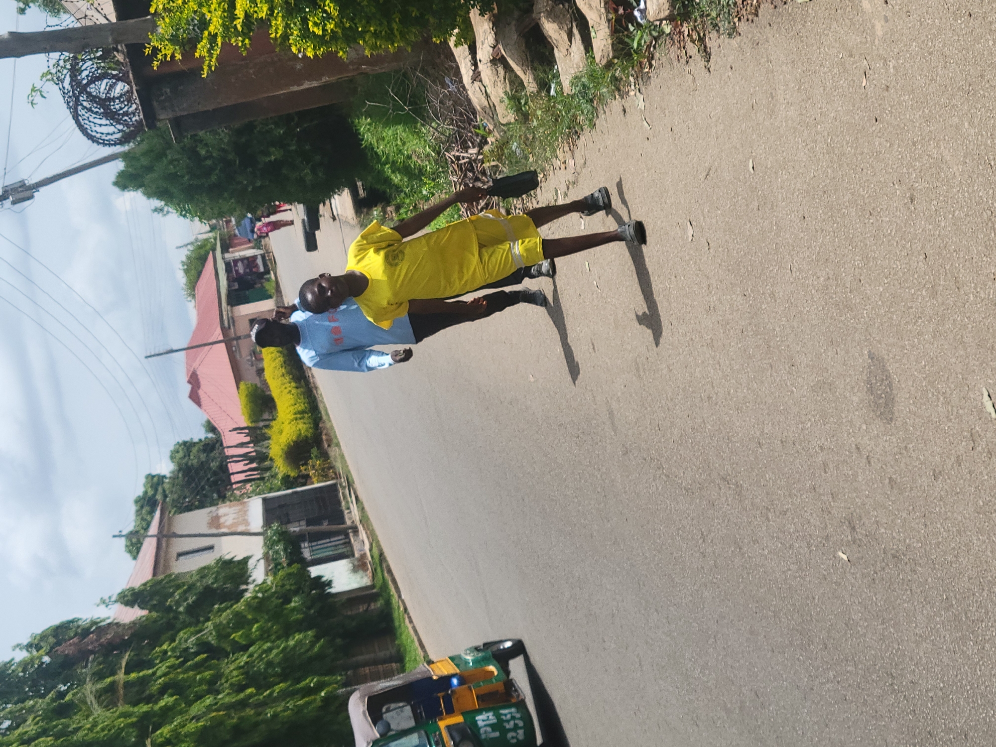 Two young men are standing on a paved road in Jos, Nigeria. One wears a yellow tracksuit and dark shoes, the other a light blue long-sleeved shirt and dark pants. The man in yellow holds a dark, rectangular object. The road is lined with buildings and vegetation. Overhead, there is a utility pole with coiled barbed wire. A green and yellow vehicle is parked at the bottom of the image.