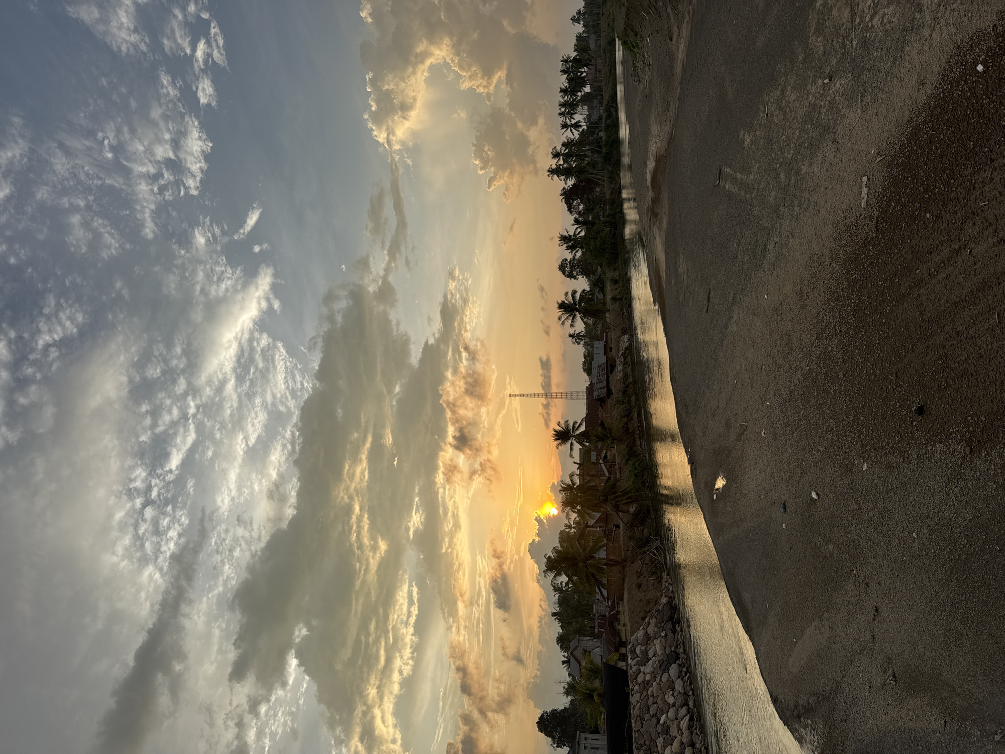 The image depicts a serene sunset scene in Meunasah Pupu, Indonesia. A body of water, possibly a river or canal, flows through the foreground, reflecting the warm hues of the setting sun. The water's edge transitions into a sandy or gravelly embankment on the right, dotted with scattered debris. Across the water, a village is visible, characterized by a cluster of houses with roofs of various colors, interspersed with numerous palm trees. A tall communication tower stands prominently on the right side of the village. The sky above is a spectacle of dramatic clouds, painted with shades of orange, yellow, and gray, with the sun partially obscured by clouds as it descends. The overall impression is one of tranquil rural life bathed in the golden light of dusk.