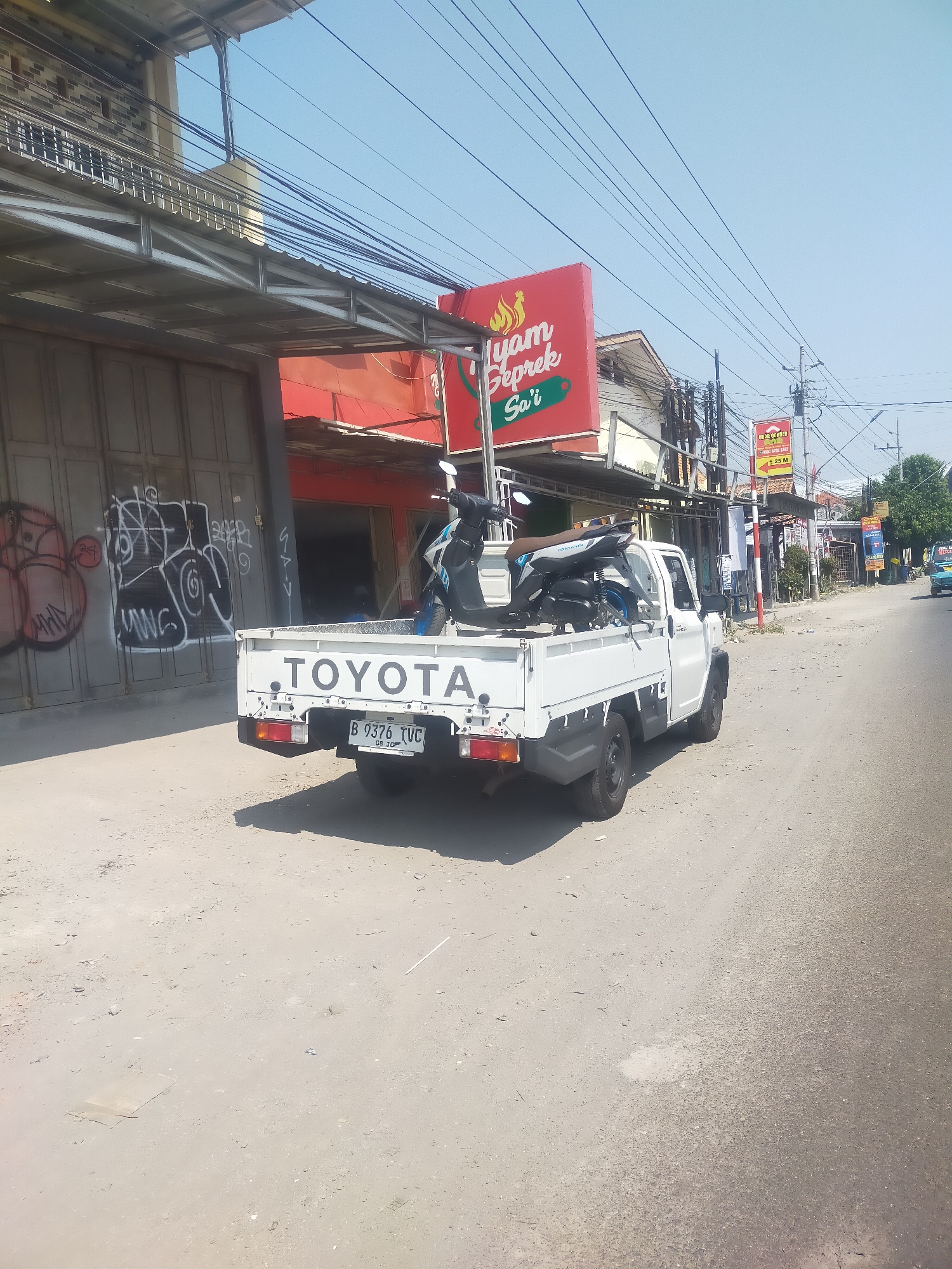 The image depicts a white Toyota pickup truck parked on a dirt road in Talang, Indonesia. The truck is carrying two motorcycles in its bed. In the background, there are several buildings with corrugated metal roofs and awnings, some of which display signage. One prominent sign is red and reads "Iyam Geprek Sari." There is graffiti on a large gray metal door to the left of the truck. Power lines crisscross overhead. The sky is clear and blue. The road surface appears to be a mixture of dirt and asphalt.