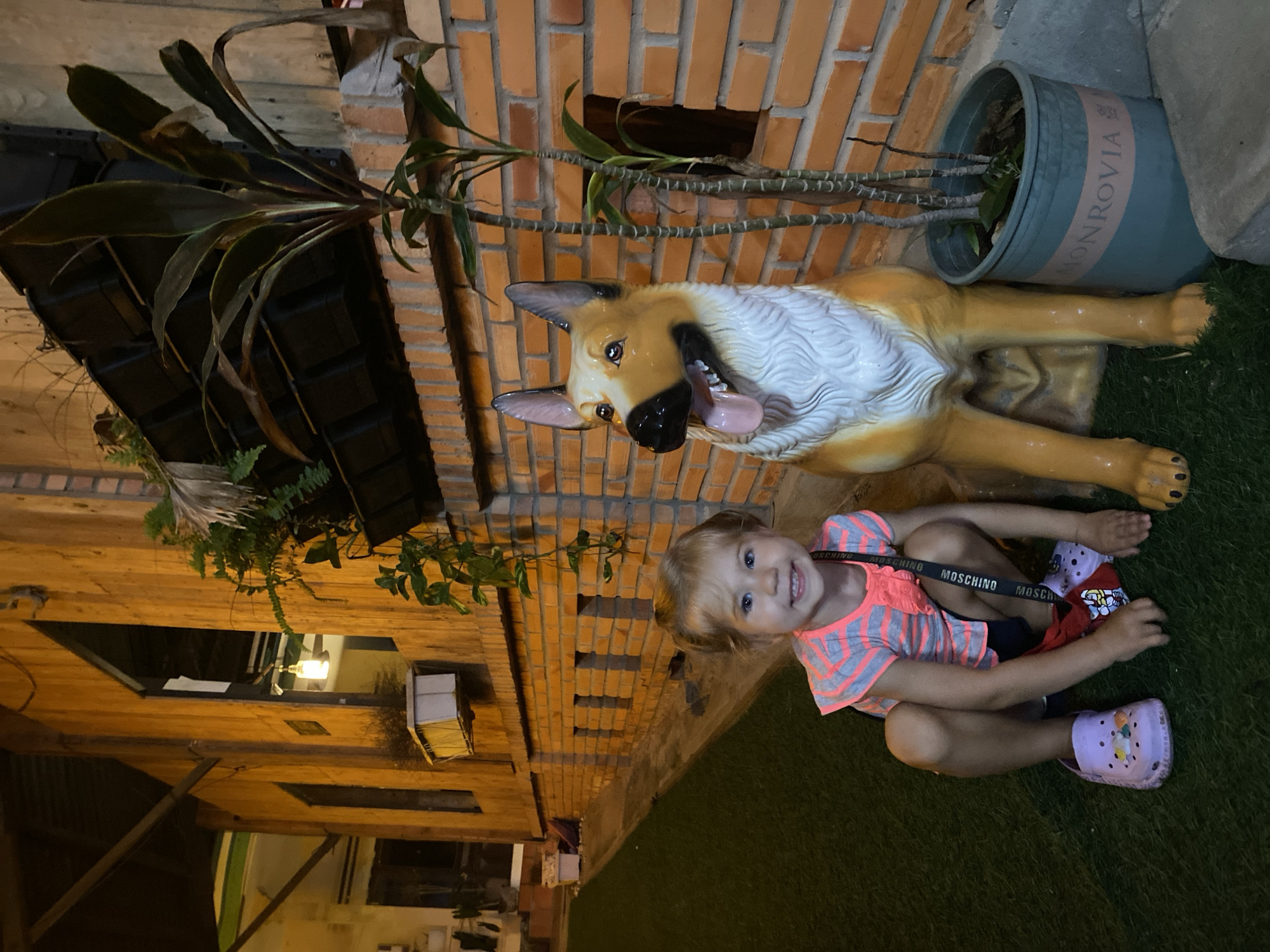 A young Caucasian girl with blonde hair and blue eyes is crouched on artificial grass in front of a statue of a German Shepherd dog. She is wearing a pink and gray striped t-shirt with ruffles on the sleeves, dark shorts, and purple crocs adorned with colorful charms. She is smiling at the camera and has a black lanyard around her neck. The dog statue is life-sized and appears to be made of ceramic or resin, with its mouth open and tongue hanging out. To the right of the dog statue is a potted plant with several green stalks and leaves. Behind them is a brick wall. The background consists of a wooden building with large windows and hanging plants. The scene appears to be outdoors in a warm climate, possibly a courtyard or walkway. The setting could be a restaurant, cafe, or tourist attraction in Mũi Né, Vietnam, given the architectural style and tropical foliage.