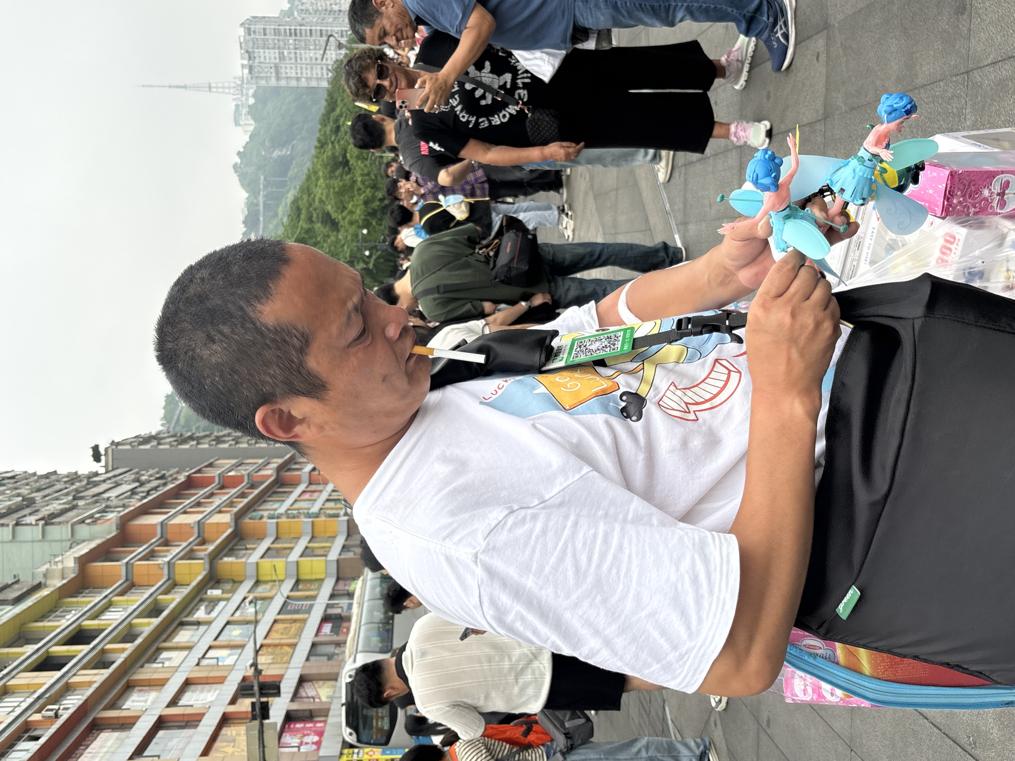A street-level, medium shot shows a middle-aged Asian man holding two small, plastic figures with blue hair and wings. He has a cigarette in his mouth and is wearing a white t-shirt with a cartoon graphic, a black bag, and an identification tag. He appears to be inspecting or assembling the figures. Behind him, a crowd of people is visible, some wearing face masks and using mobile phones. In the background, a colorful, multi-story building with repeating window patterns stands out. A street and vehicles are partly visible at the lower edge of the image. The scene takes place in the city of Yuzhong District, China.