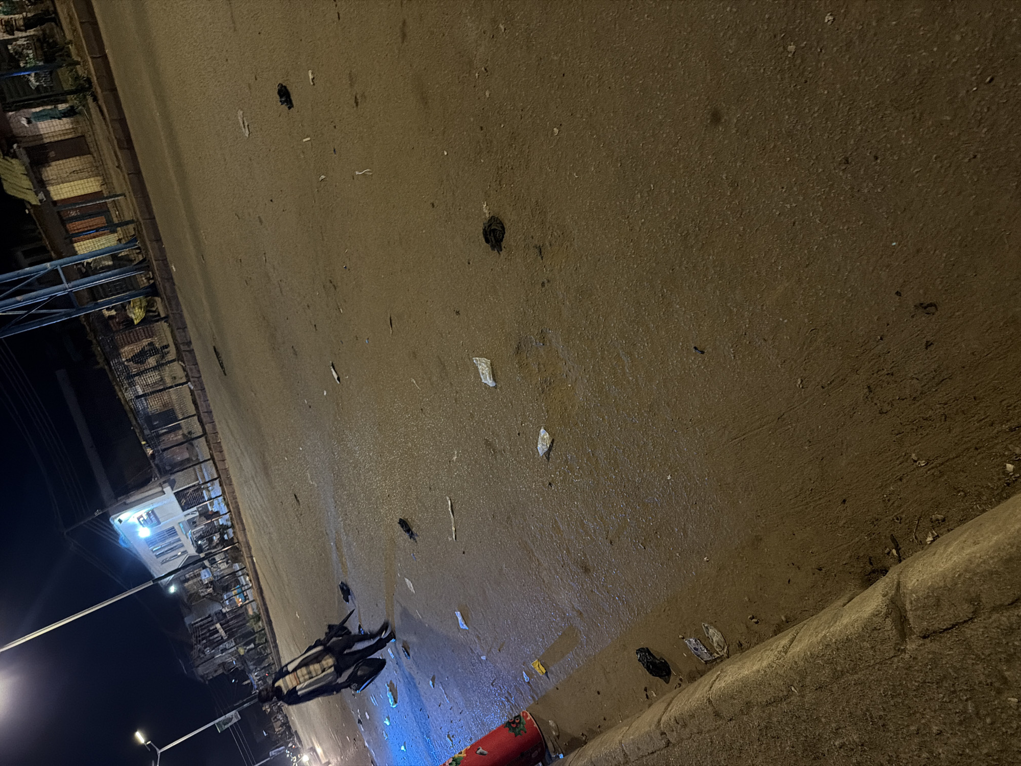 In Bukuru, Nigeria, a night scene unfolds on a street littered with debris. The road surface appears wet, reflecting the artificial light from streetlamps and buildings. A person, seen from the waist down, walks across the street carrying a dark bag. The individual is wearing dark trousers and a patterned shirt. Scattered across the road are various pieces of trash, including plastic bags and paper. In the background, buildings with lit windows line the street, and a fence encloses a property. A red traffic cone with a festive pattern is visible on the left side of the image. The overall atmosphere suggests an urban environment after dark, with signs of recent activity or accumulation of waste.