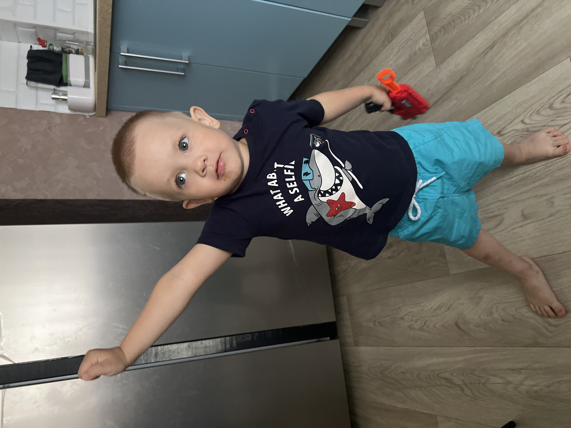 In the city of Марковский, Russia, a young fair-skinned male child with short light brown hair and blue eyes stands in a domestic kitchen environment. He is wearing a dark blue t-shirt featuring a cartoon shark with sunglasses and a red starfish bowtie, accompanied by the text "WHAT AB.T A SELFIE!". He also wears bright turquoise shorts with a white drawstring and is barefoot. His left arm is extended upwards and backward, with his hand gripping the vertical edge or handle of a large, reflective stainless steel refrigerator. His right hand holds a red and black toy, resembling a small shovel or scoop. The child's gaze is directed slightly upward and to the left. A faint smudge is visible on his upper lip. The floor beneath him consists of light-colored wooden laminate. To the right, a kitchen area is visible with teal-colored cabinets, a light brown countertop, and a white tiled backsplash. A white organizer caddy containing kitchen utensils and a dark cloth are present on the countertop.