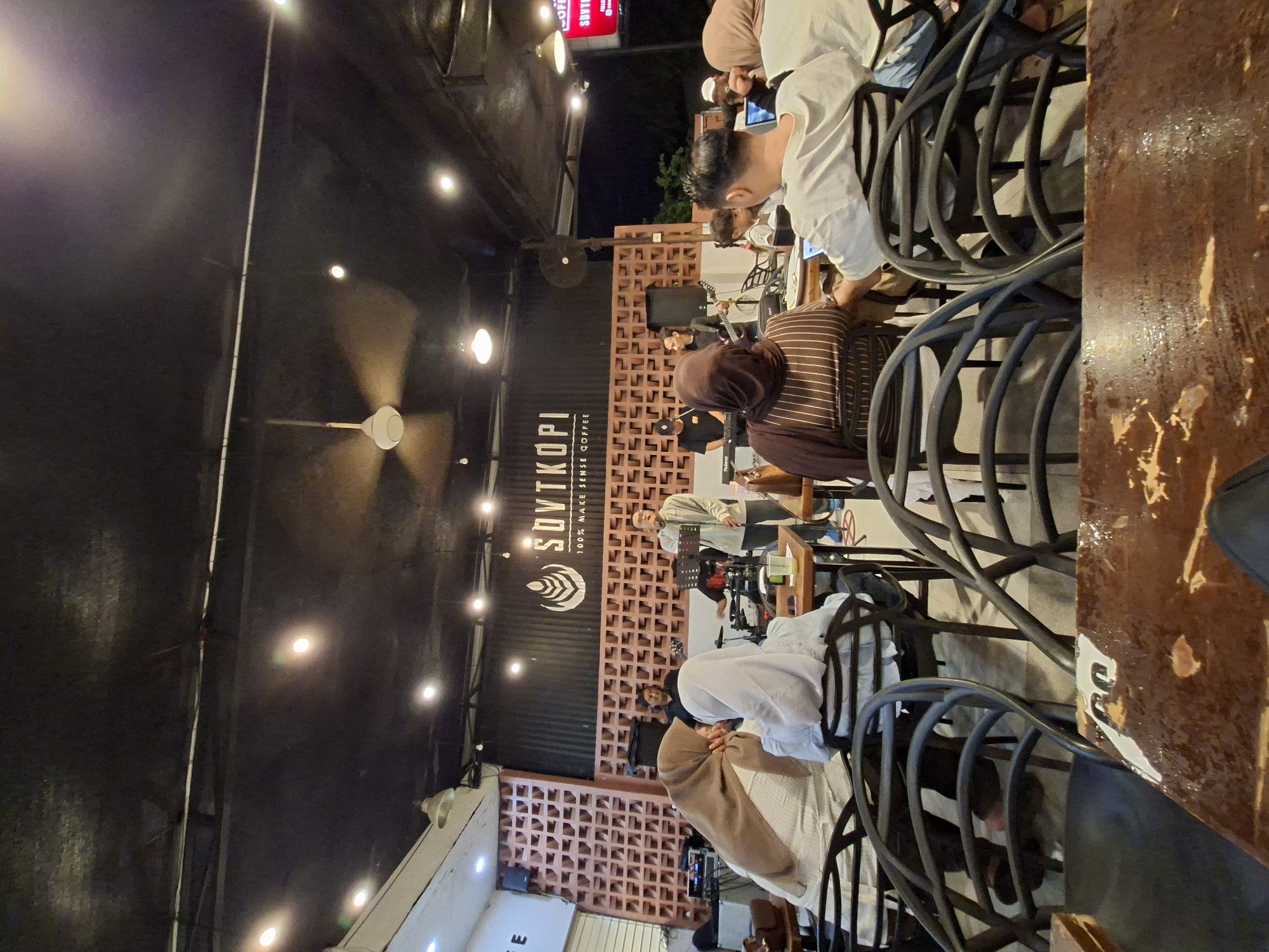 The image shows an outdoor seating area at Sovtkopi, a coffee shop in Lhokseumawe, Indonesia. Several people are seated at tables, some are wearing head coverings and others are not. A ceiling fan spins overhead. A brick wall with a cutout design is visible behind the patrons. A band is performing in the background. The light appears to be dim and artificial, with many string lights illuminating the space.